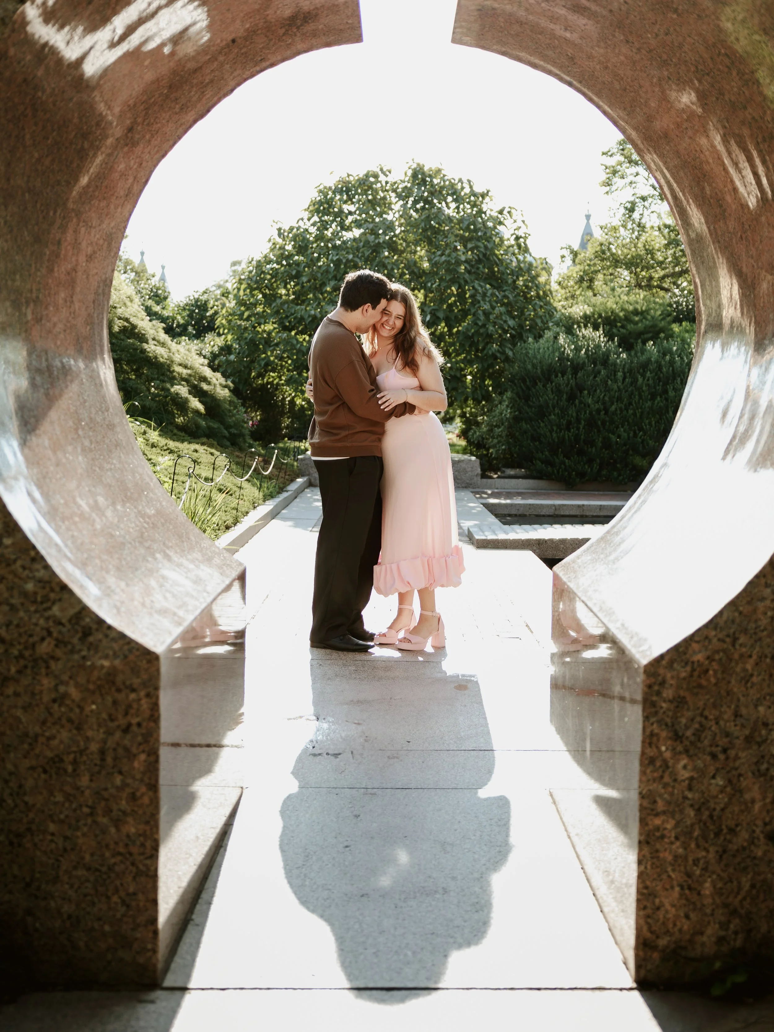 A couple standing close together, looking at each other and smiling, seen through a circular opening in a sculpture or structure with greenery and trees in the background.