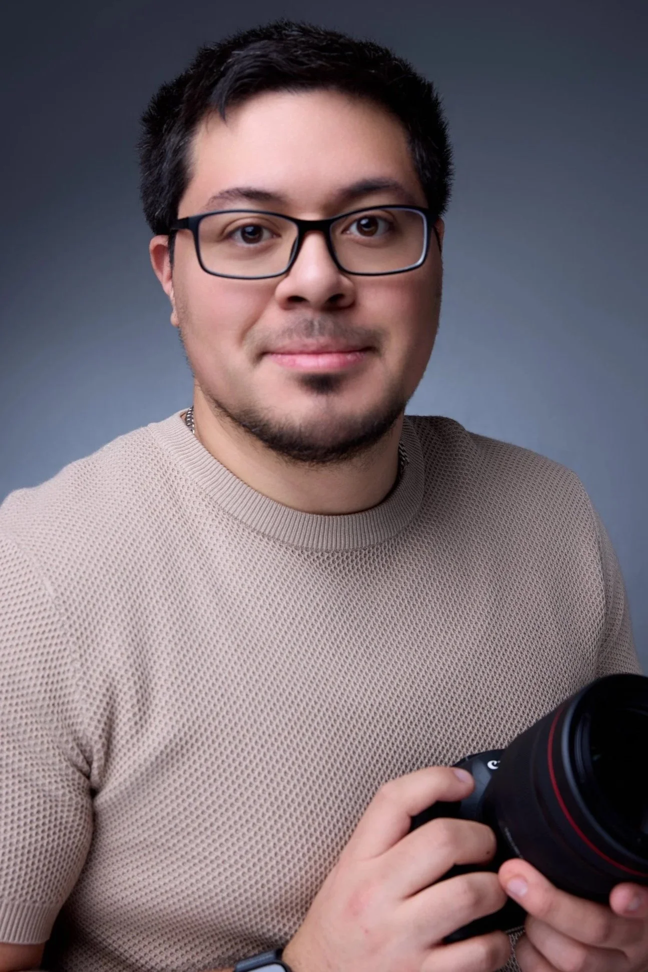 A man with glasses and a beige textured shirt holding a professional camera, smiling at the camera against a plain gray background.