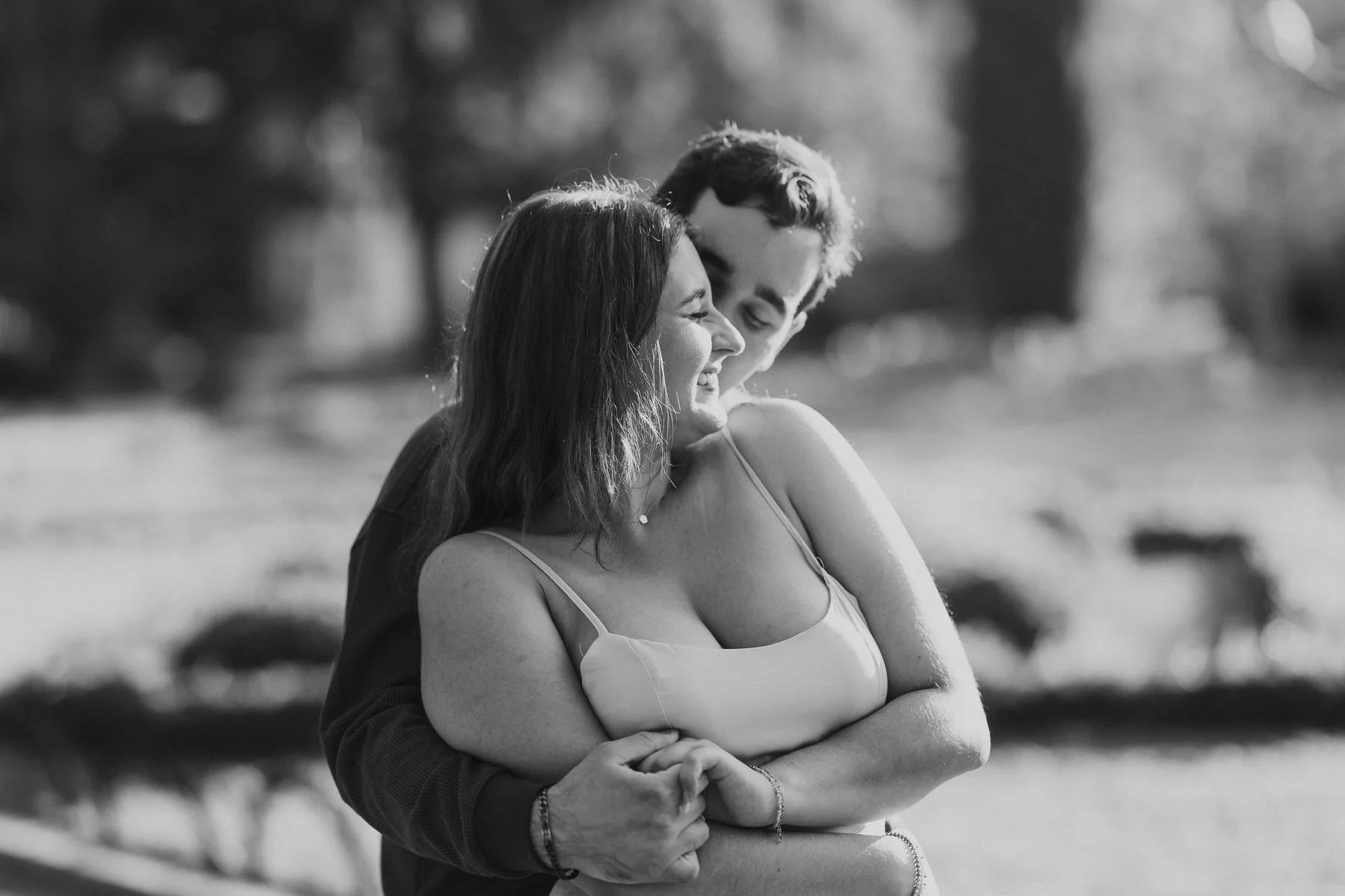 A black and white photo of a young couple smiling and hugging outdoors, with trees and blurred background.