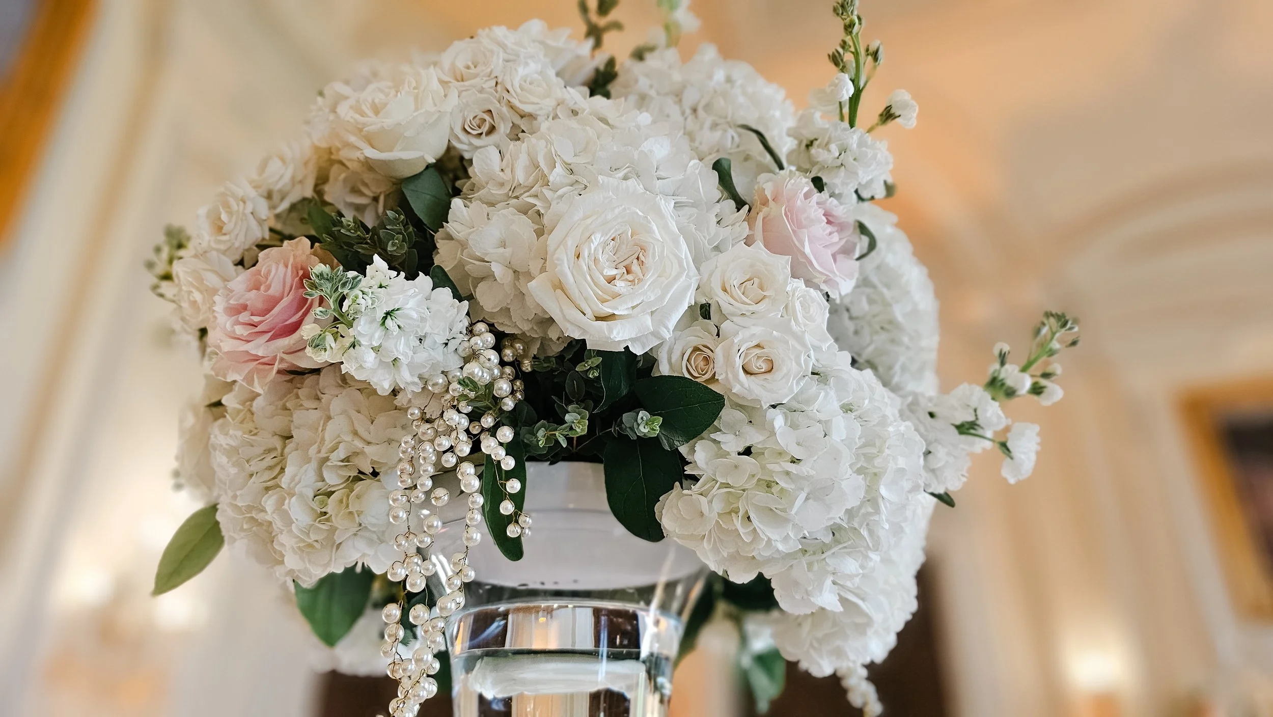 A close-up of a floral arrangement with white hydrangeas, roses, and pink flowers, accented with green leaves and pearl-like beads in a clear glass vase.
