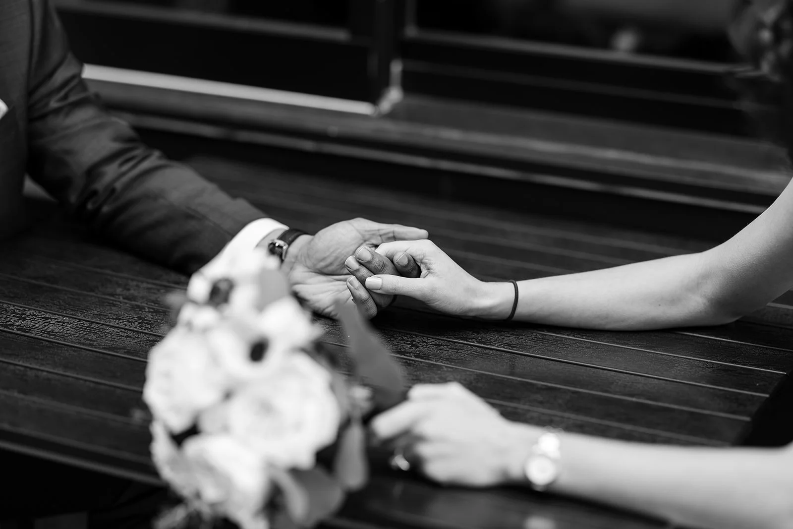 A person with long hair wearing a black wristband offers their hand to another person during a wedding ceremony, with a bouquet of flowers and a wooden table in the foreground.