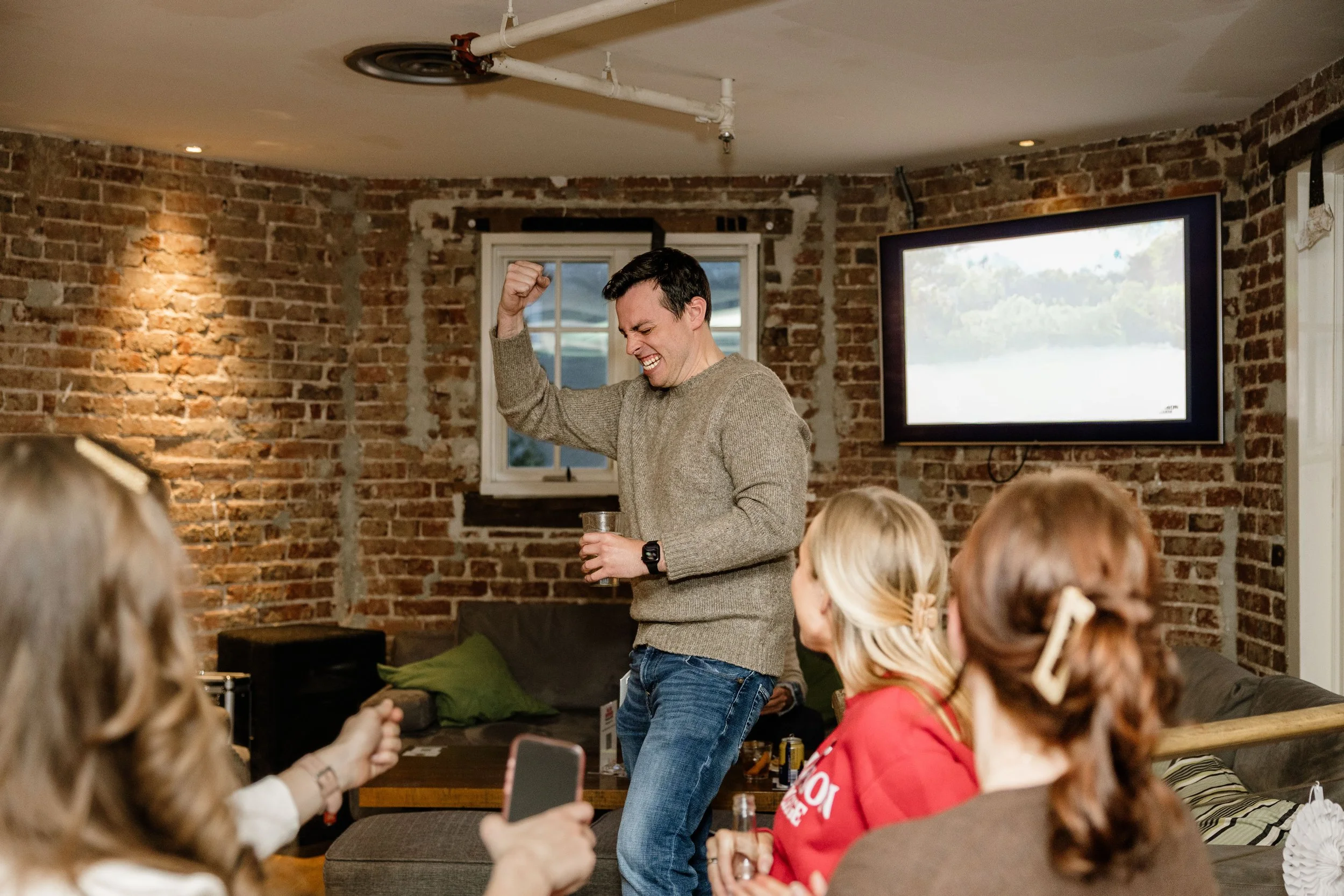 Man celebrating with a drink in hand at a casual indoor gathering with friends, brick wall background, TV on the wall.