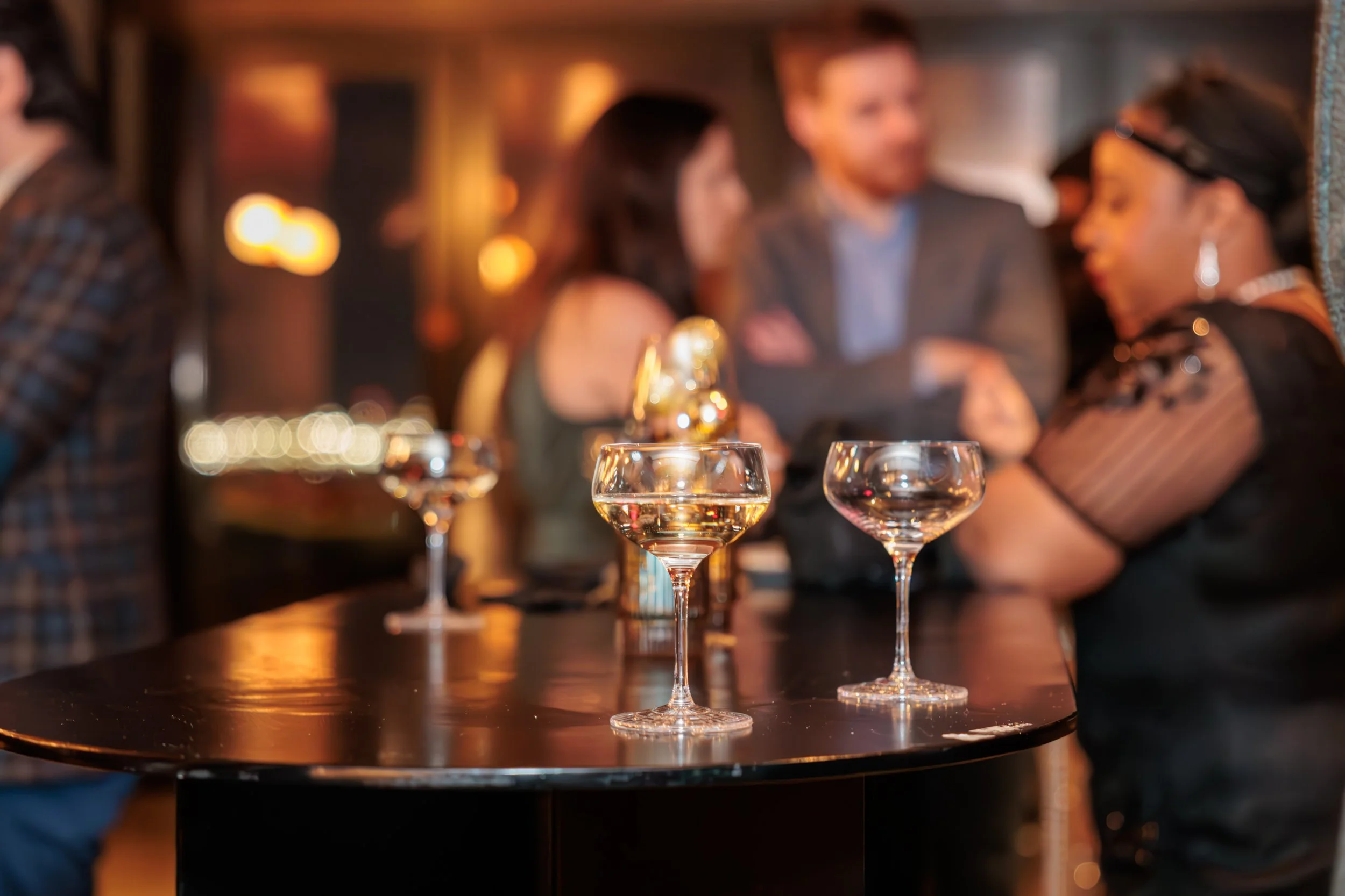 Four empty cocktail glasses on a black table, with a blurred background of people socializing in a dimly lit bar or restaurant.