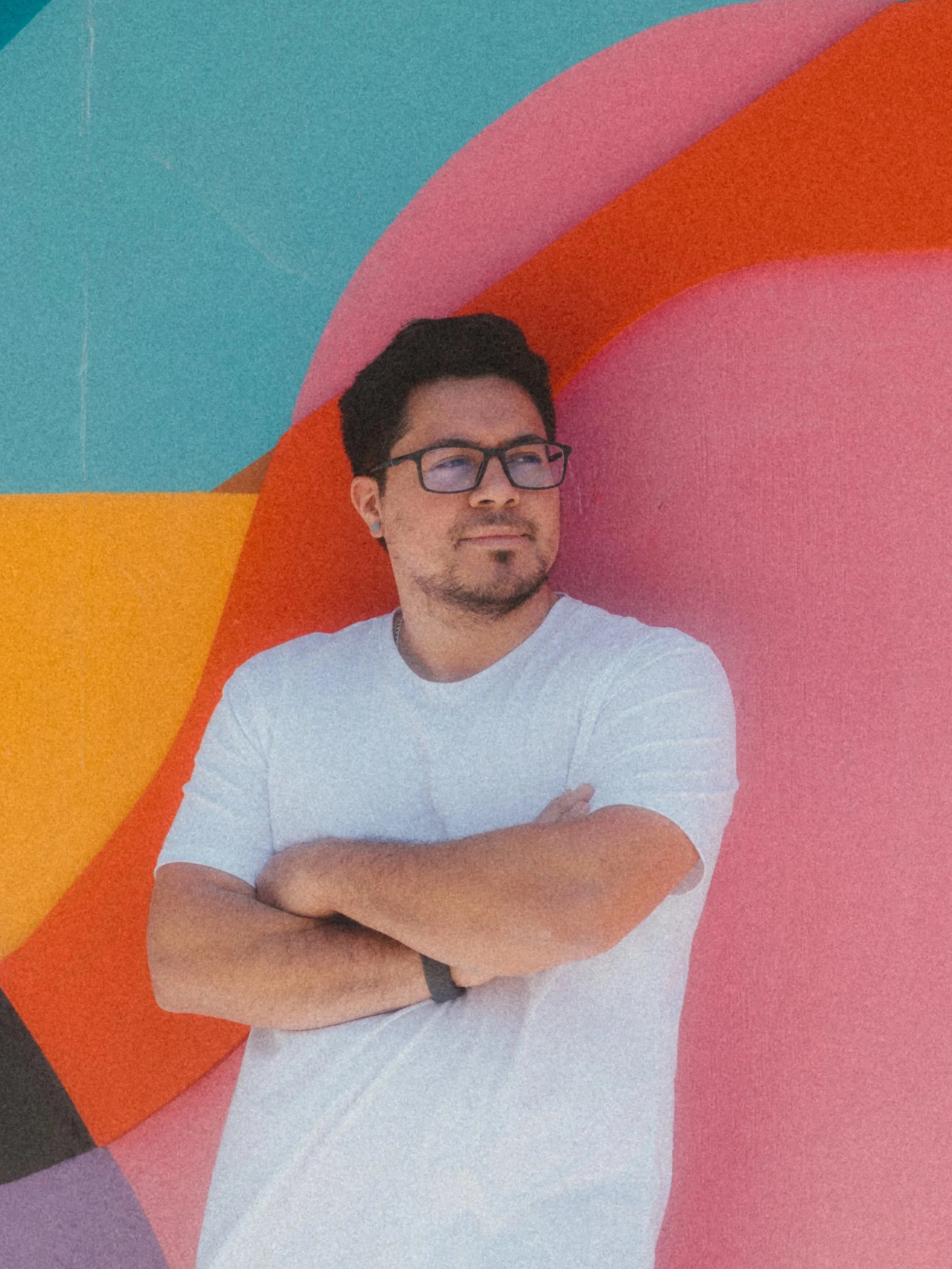 Man with dark hair and glasses standing in front of a colorful mural with pink, orange, purple, and teal sections, arms crossed, looking thoughtfully to the side.