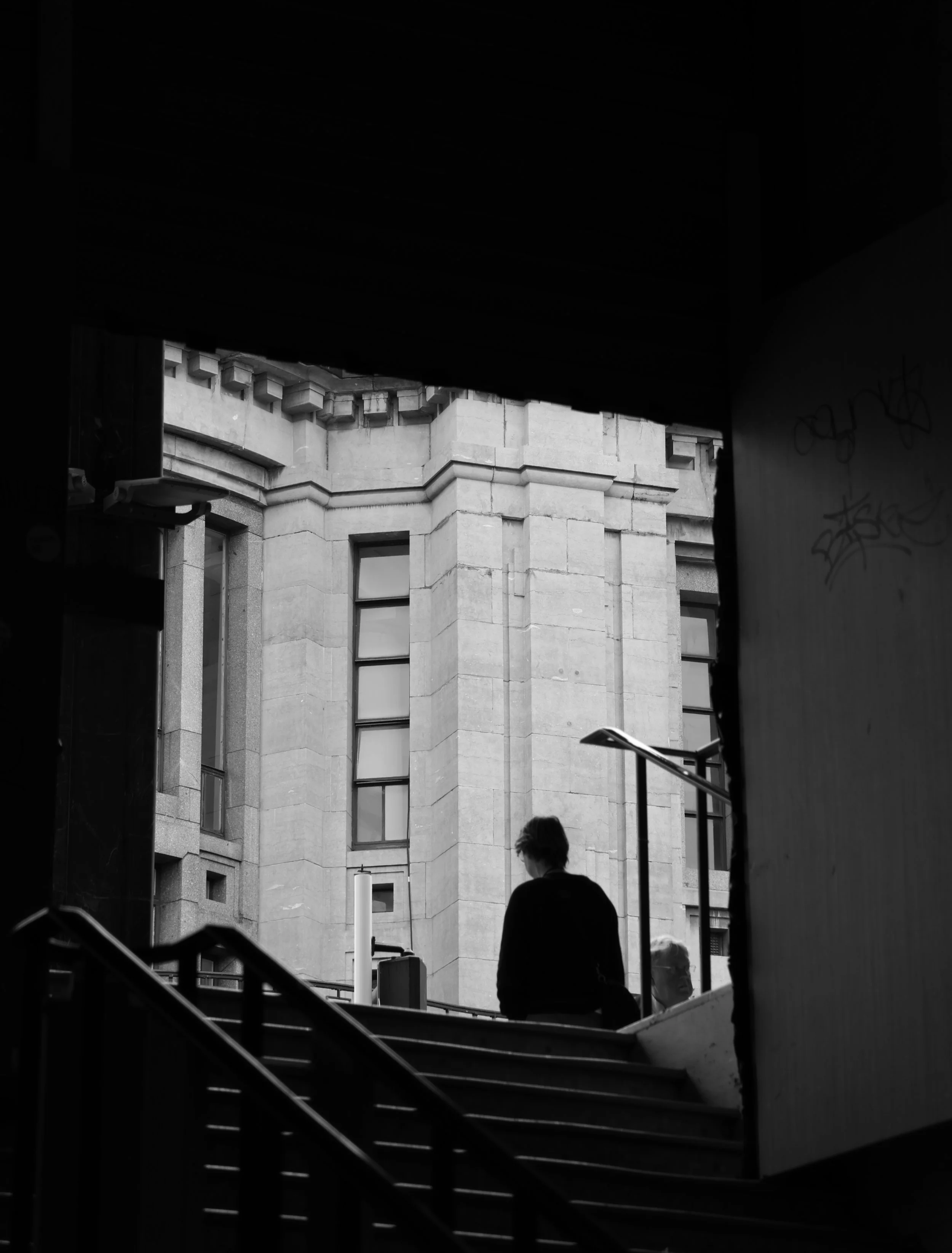 Une silhouette d'homme assis à un escalier dans une zone sombre avec un bâtiment en pierre en arrière-plan, vue en noir et blanc.
