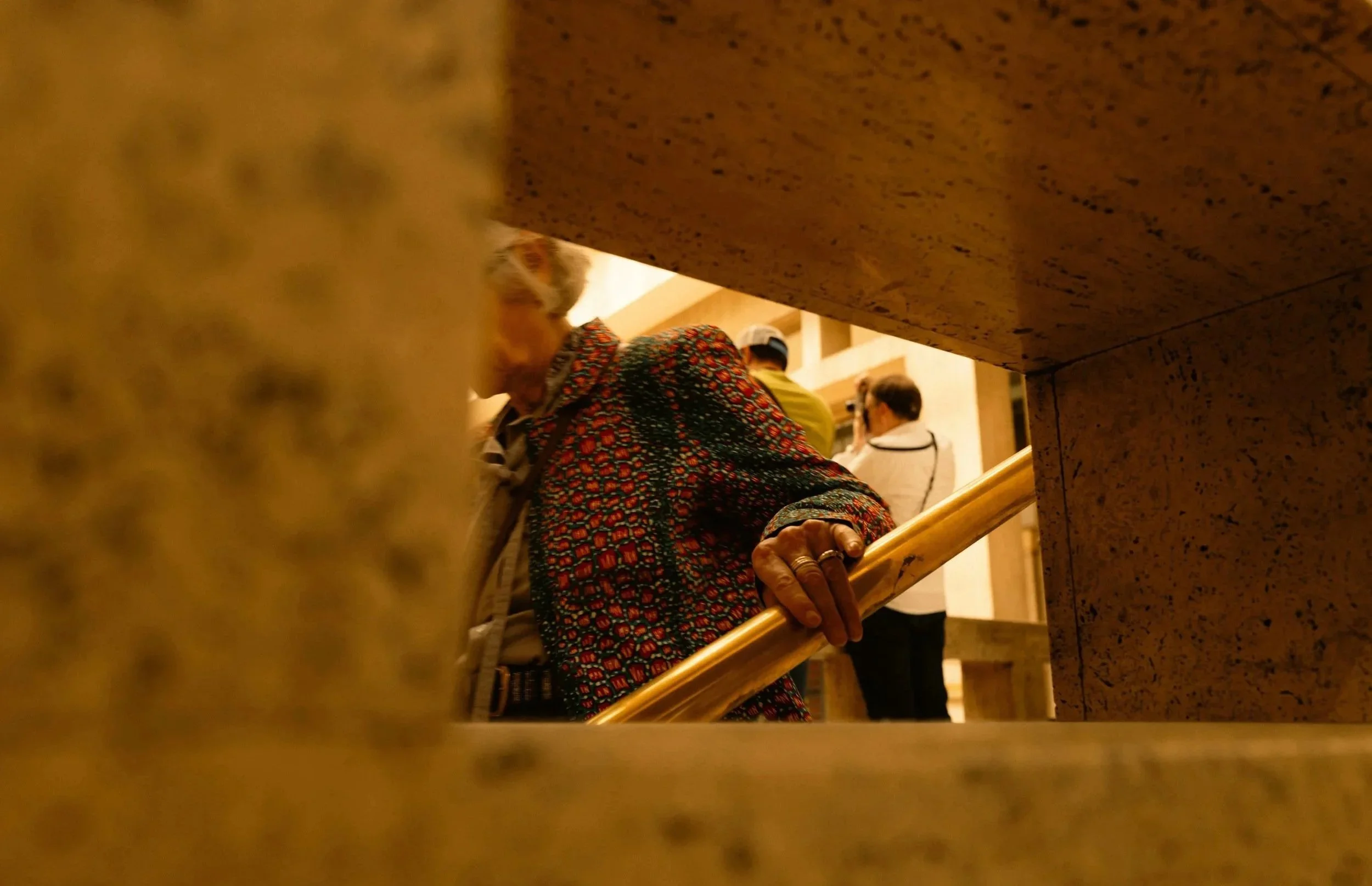 Personne âgée montant un escalier, vue partiellement cachée par une rambarde en bois, dans un bâtiment avec une ambiance chaleureuse.