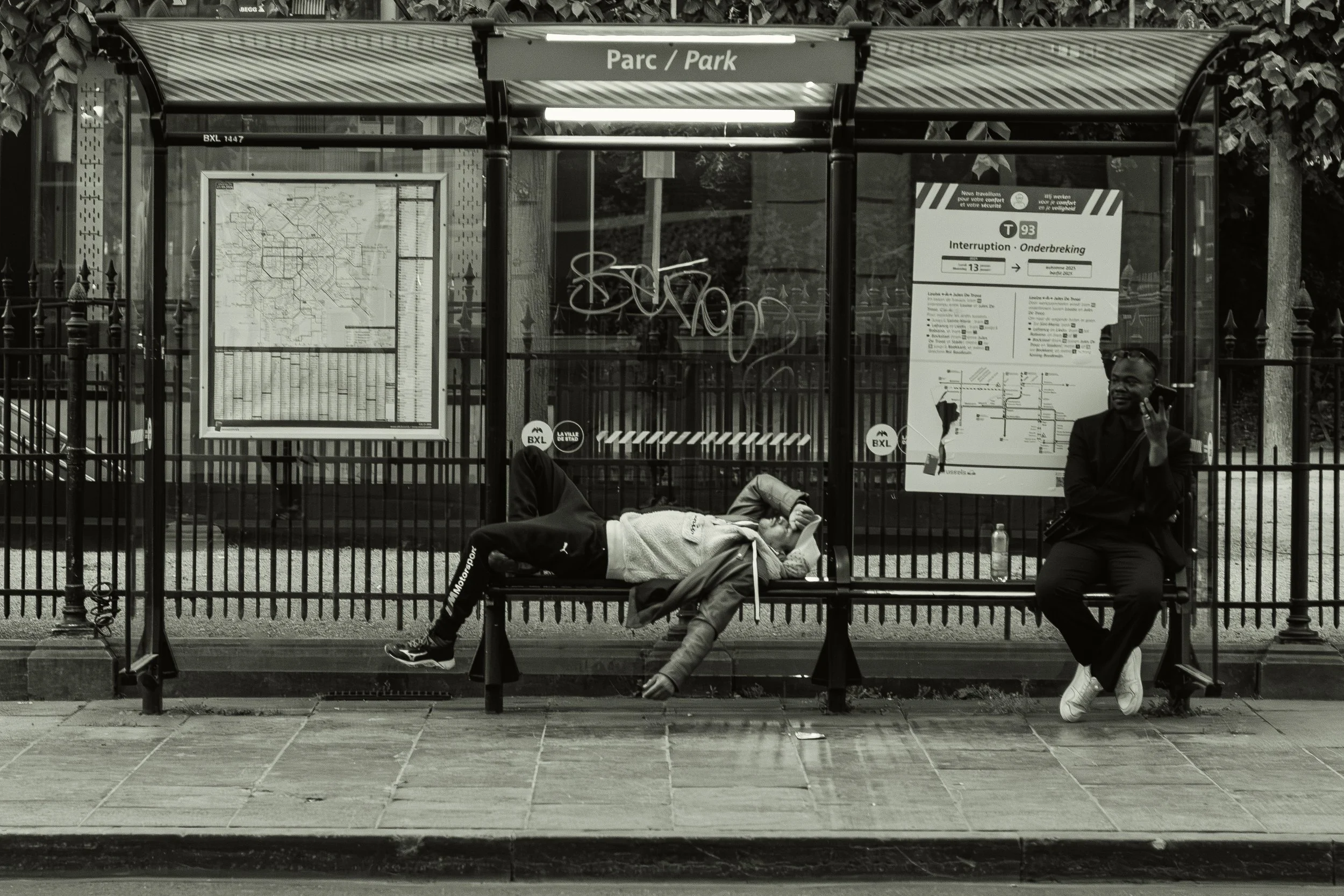 Deux personnes assises à un arrêt de bus, l'une allongée sur le banc et l'autre assise, en train de parler au téléphone, dans un parc à Bruxelles.