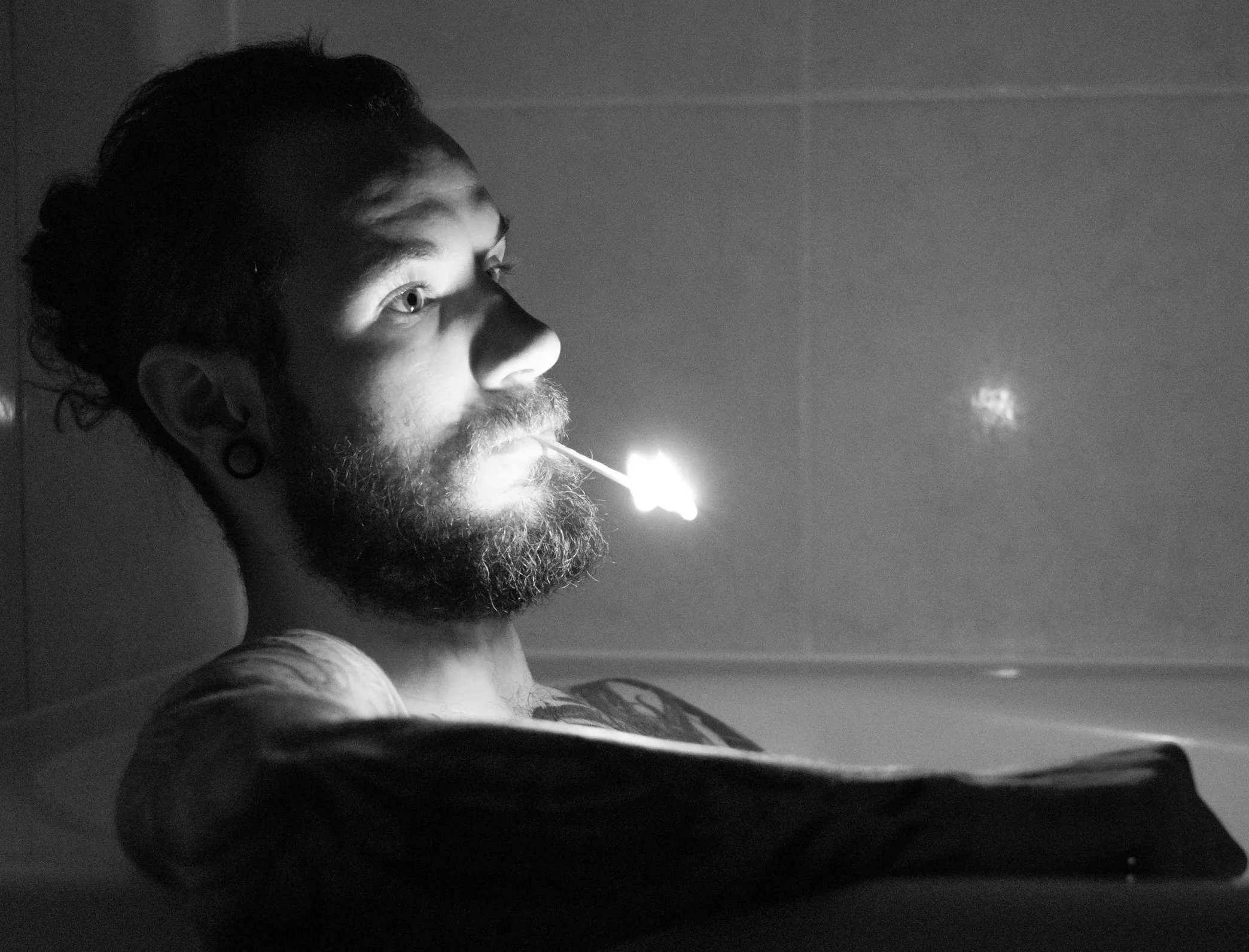 Un homme à la barbe et aux cheveux longs, assis dans un bain, tient un bâton d'encens avec de la fumée qui s'élève, éclairé par une lumière douce.