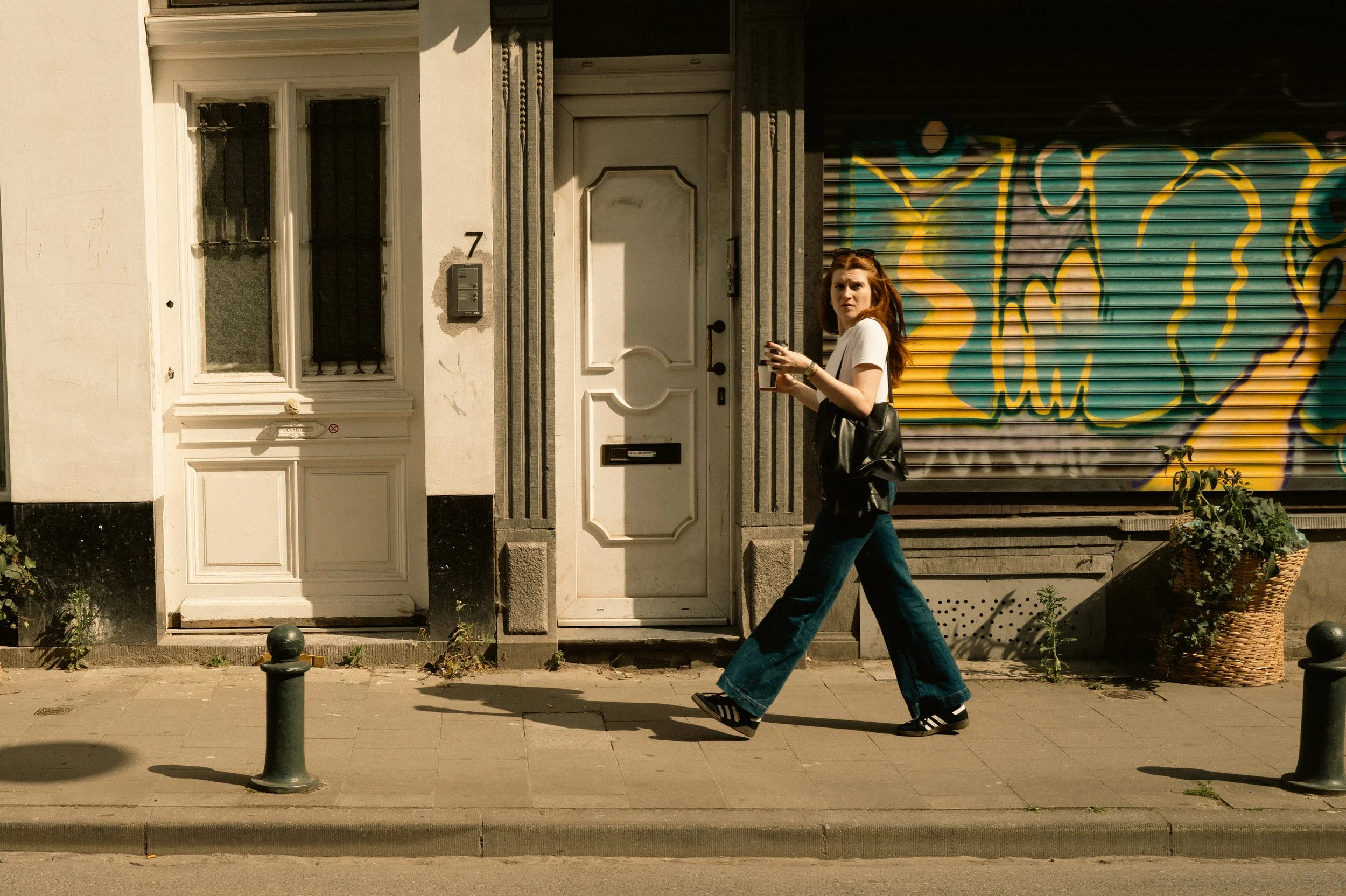 Jeune femme à la marche, portant un t-shirt blanc, un sac noir, des jeans amples, et des chaussures de sport, traversant un trottoir urbain sous un soleil brillant, avec un mur coloré en arrière-plan.