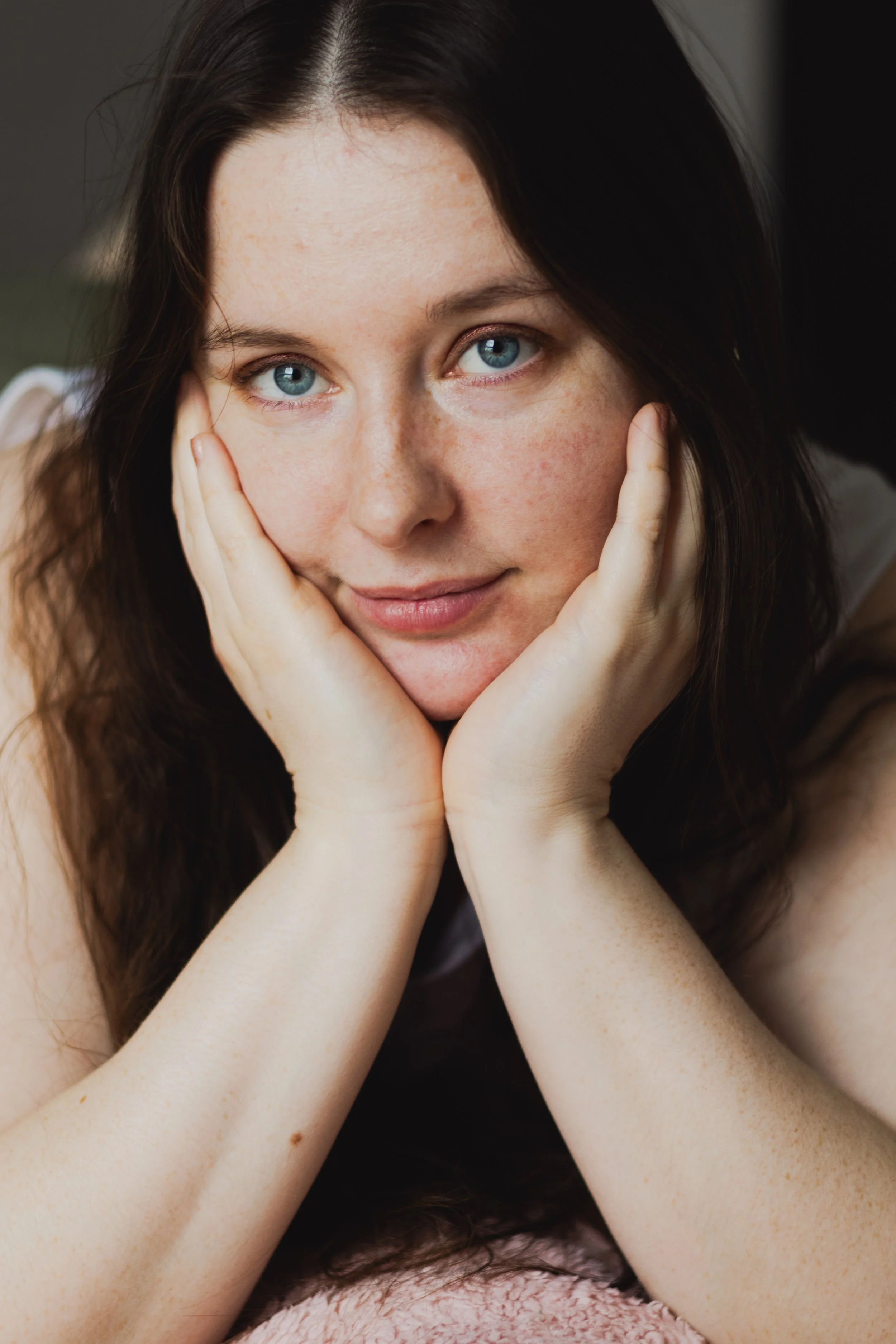 Portrait d'une jeune femme avec des yeux bleus, regardant l'appareil, les mains soutenant son visage, cheveux bruns.