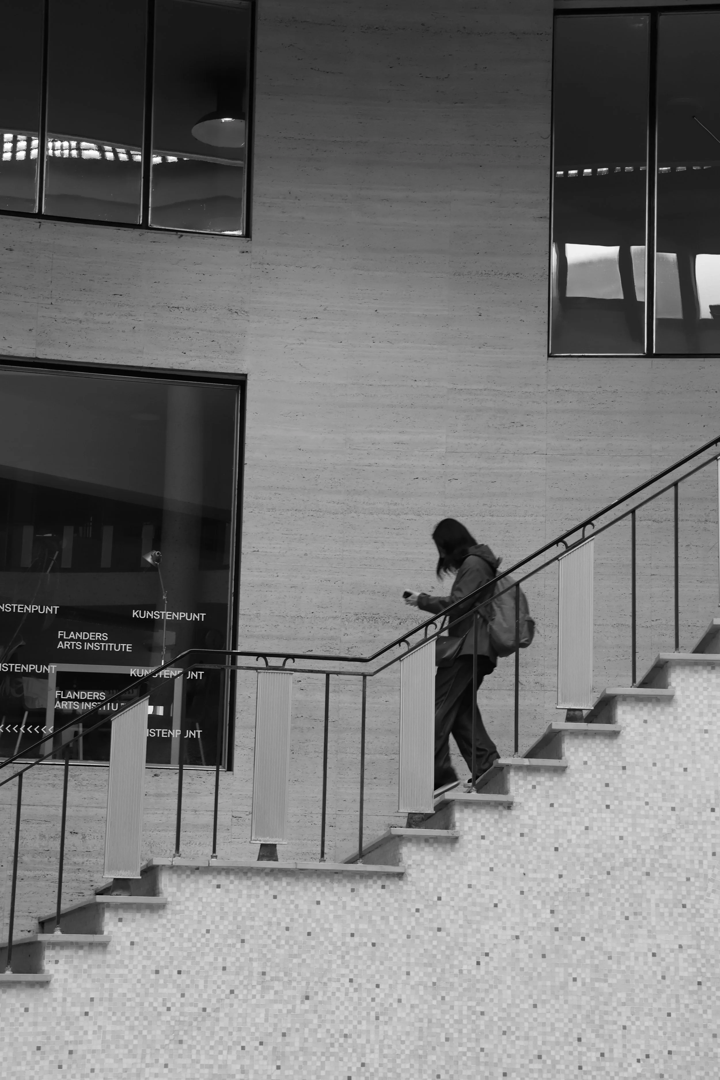 Une personne avec un sac à dos descendant un escalier dans un bâtiment moderne en bâchecies de verre et pierre, en noir et blanc.