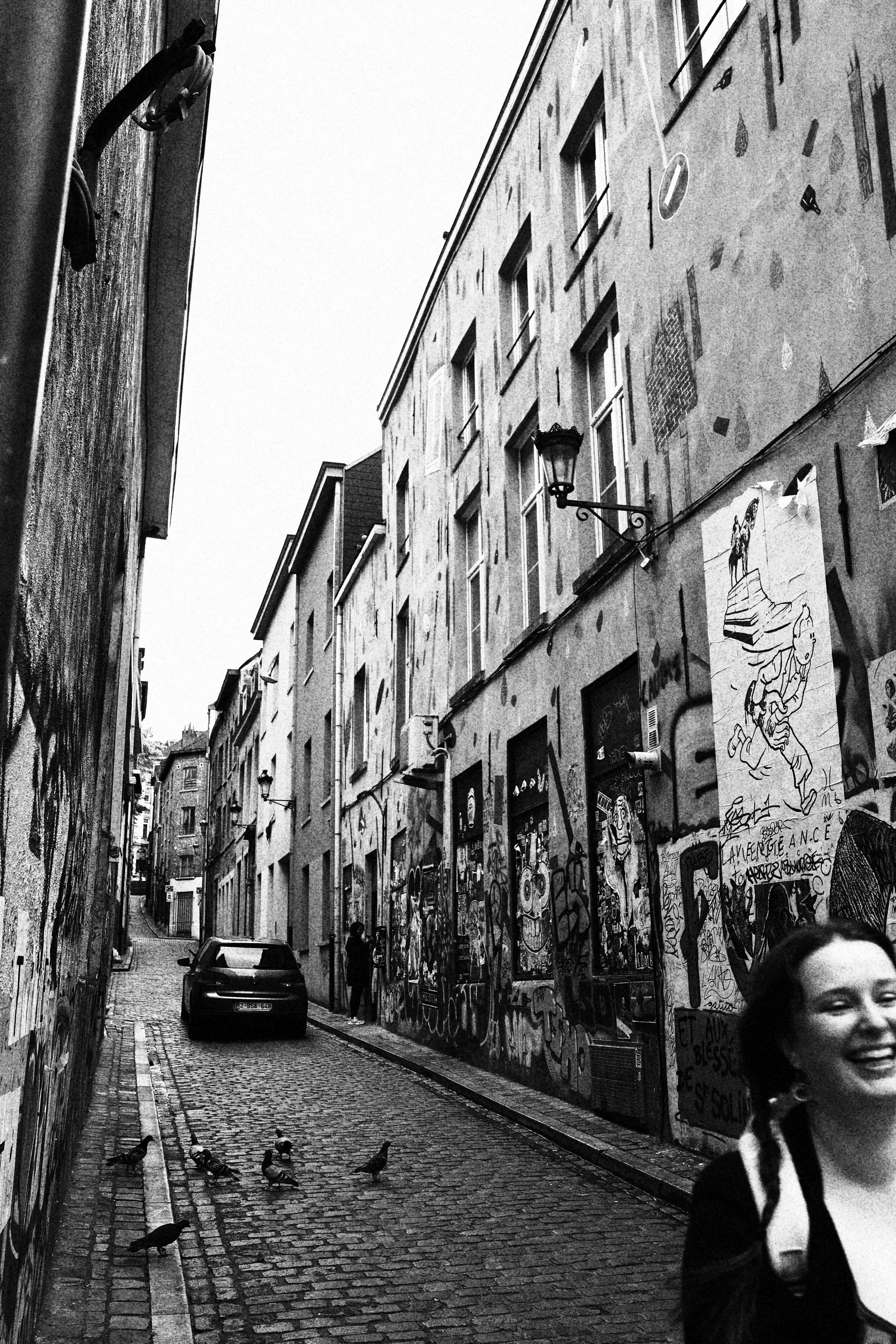 Ruelle pavée en ville avec murs graffités, un voiture stationnée, deux personnes dont une qui prend une photo, pigeons au sol, bâtiments à plusieurs étages, lampes extérieures.