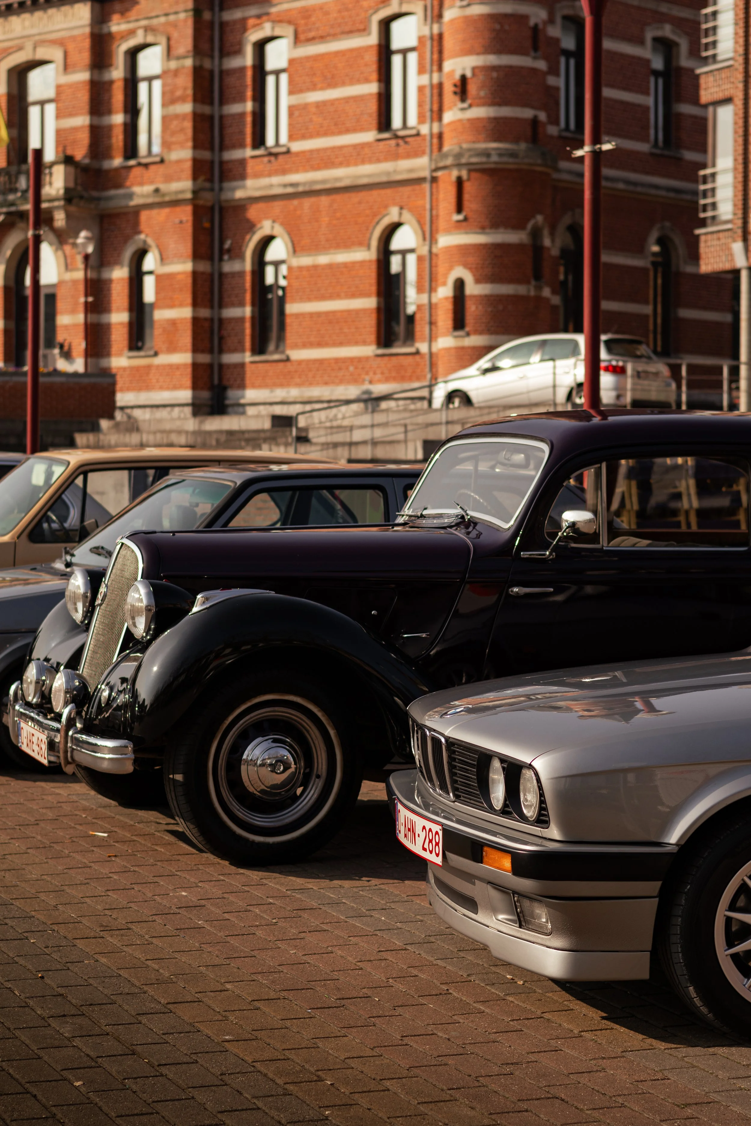 Voitures classiques stationnées devant un bâtiment en briques rouges avec de grandes fenêtres.