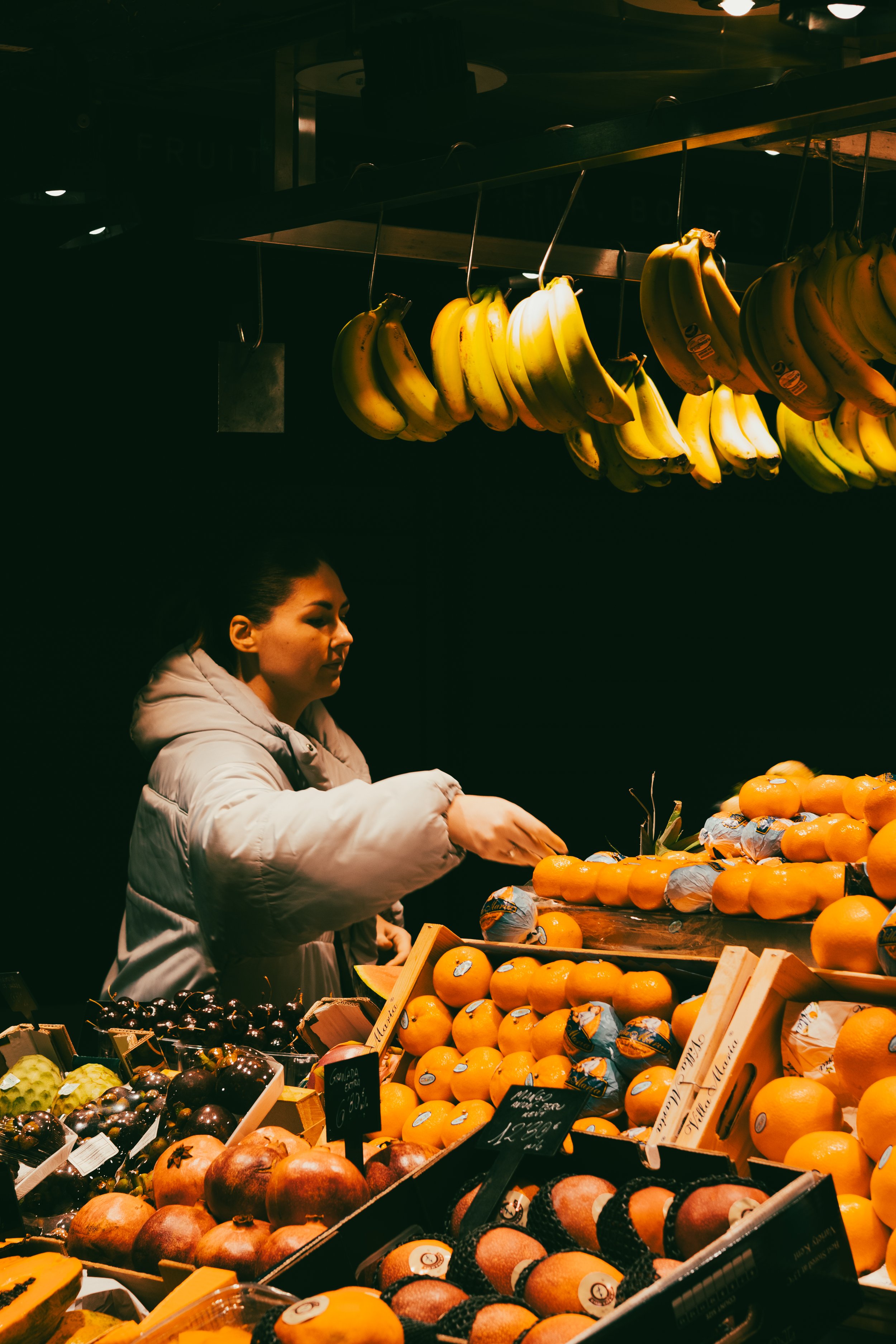 Femme achetant des agrumes dans un stand de marché nocturne avec des bananes suspendues au plafond.