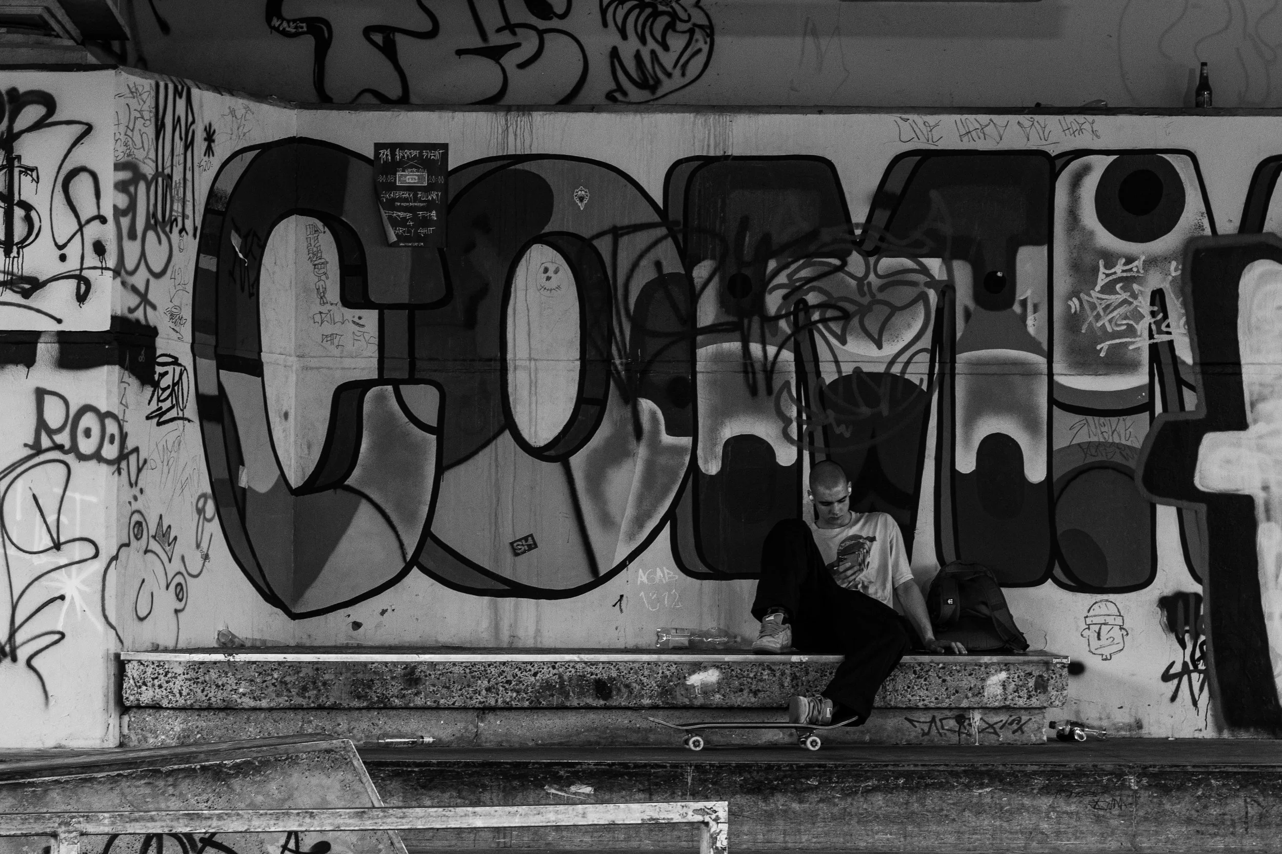 Jeune homme assis sur un banc en dessous d'un graffiti urbain, café à la main, dans un environnement de skatepark ou de sous-sol graffité.