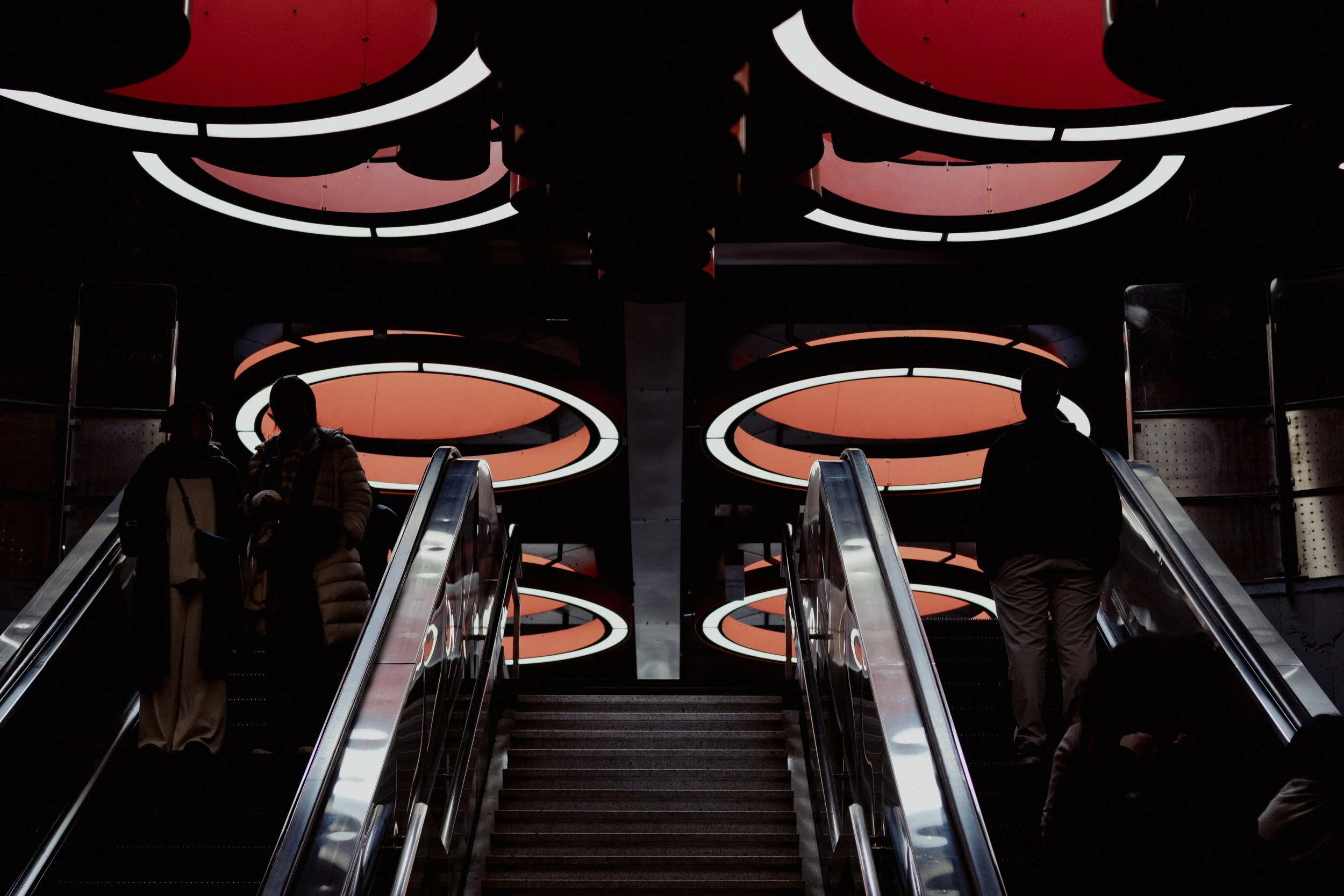 Silhouettes de personnes descendant des escalators dans un bâtiment moderne avec un plafond lumineux orange et circulaire.
