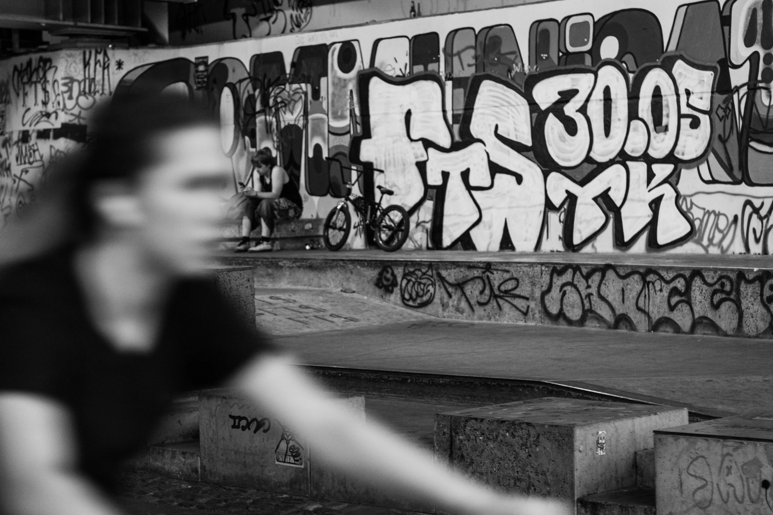 Une scène de graffiti urbain avec un jeune homme assis sur un banc, une bicyclette et un mur couvert d'art urbain en arrière-plan, ainsi qu'une personne floue passant devant la caméra.