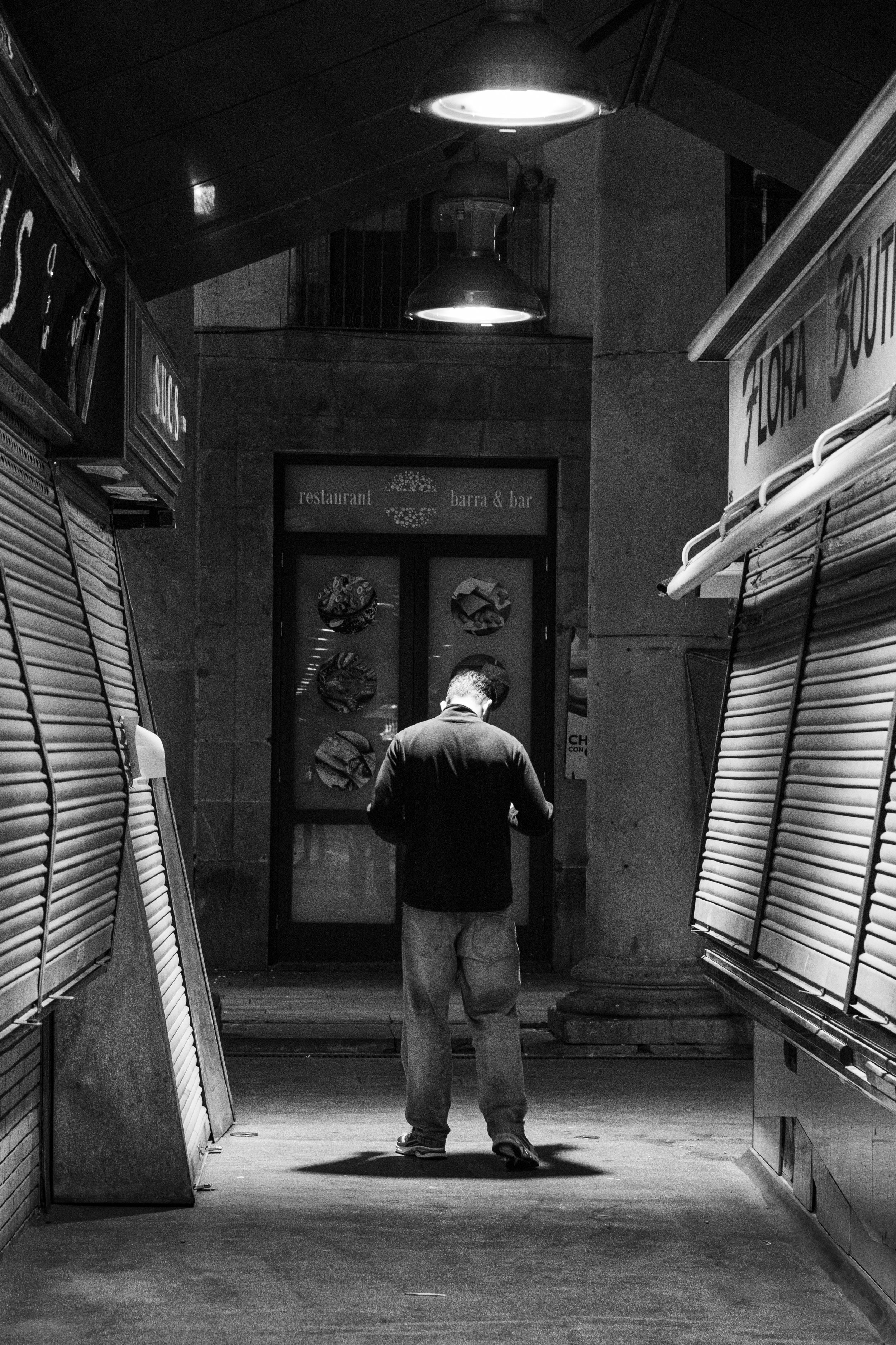 Une personne se tient devant la porte d'un restaurant ou bar en soirée, dans une rue silencieuse et éclairée par des lampes suspendues, entourée de vitrines de marchands fermés.