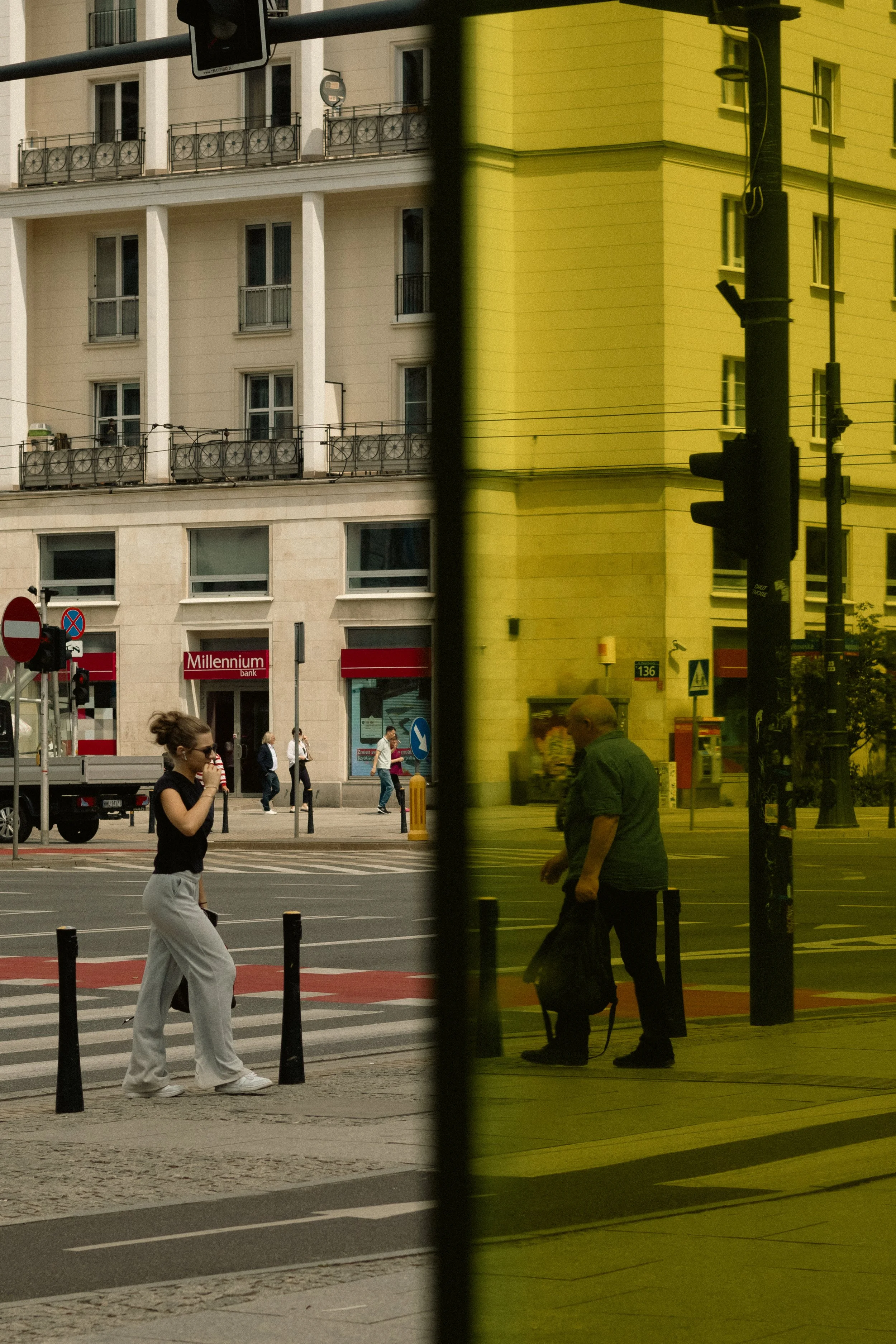 Une reflection sur une vitre montrant deux personnes traversant une rue à proximité d'un bâtiment jaune. Une femme marche en portant un café et un homme se tient avec un sac.