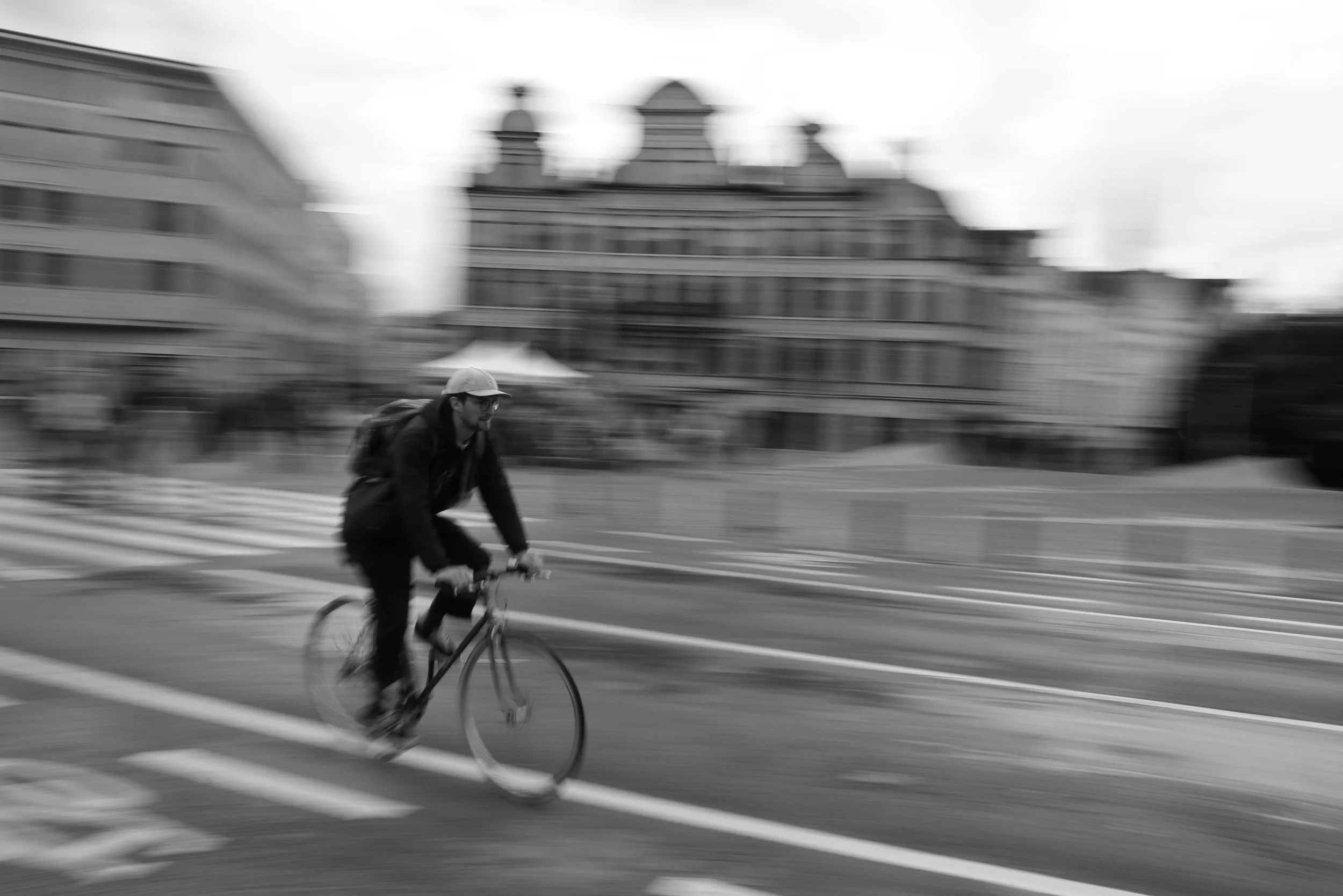 Un cycliste portant un casque et un sac à dos, roulant à vélo sur une rue urbaine. La photo est floue, indiquant le mouvement, et en noir et blanc.