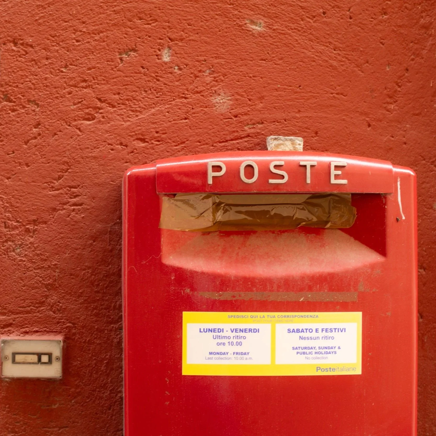 Une boîte aux lettres rouge contre un mur rouge, avec une étiquette indiquant les jours de collecte du courrier.