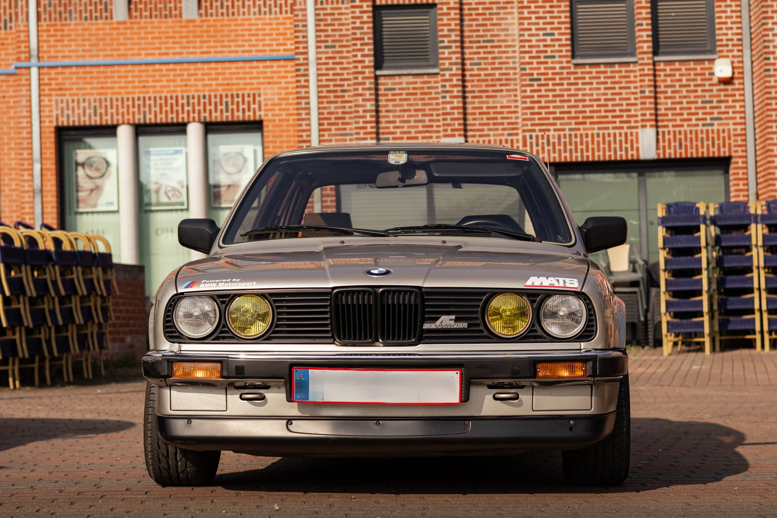 Voiture BMW classique, stationnée devant un bâtiment en briques, avec des affiches de lunettes sur une fenêtre derrière.