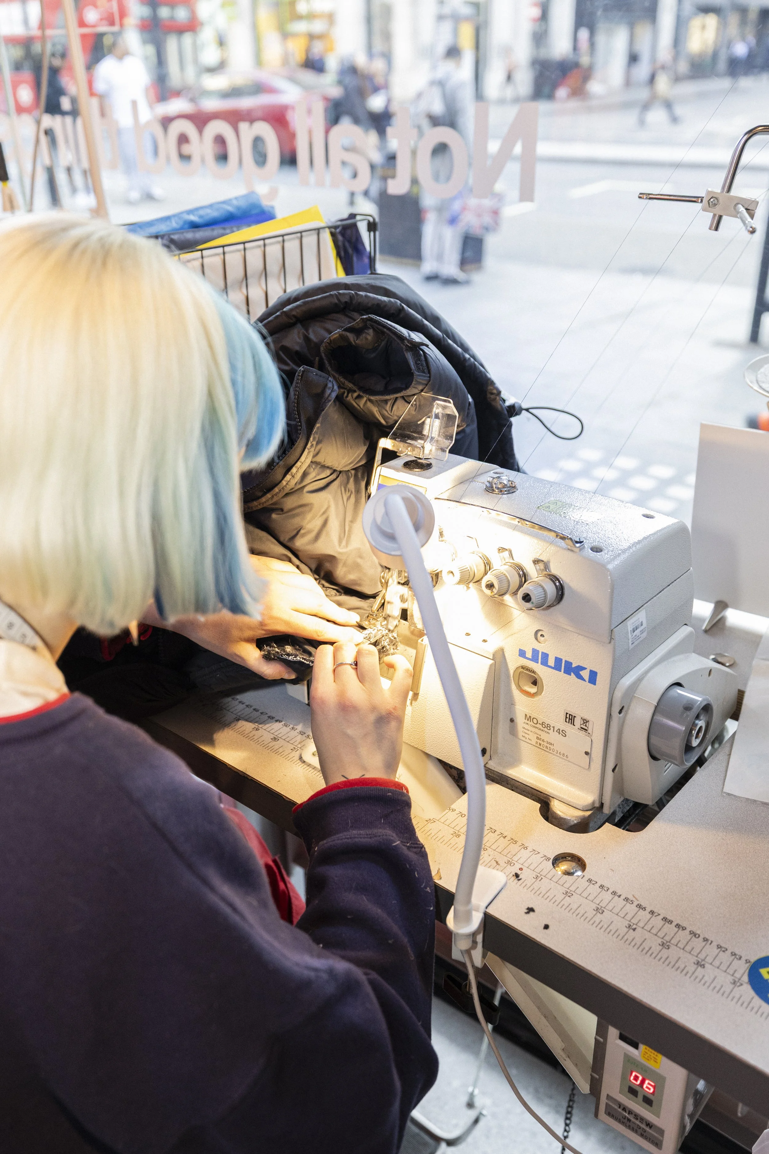 Person sewing on a JUKI industrial sewing machine in a store, with street outside visible through window.