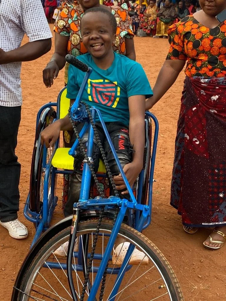 A smiling young boy in a wheelchair on a dirt field, surrounded by people. She is wearing a bright blue T-shirt with colorful graphic design, and is holding the handlebars of the blue wheelchair. The scene appears to be in an outdoor community or gathering.