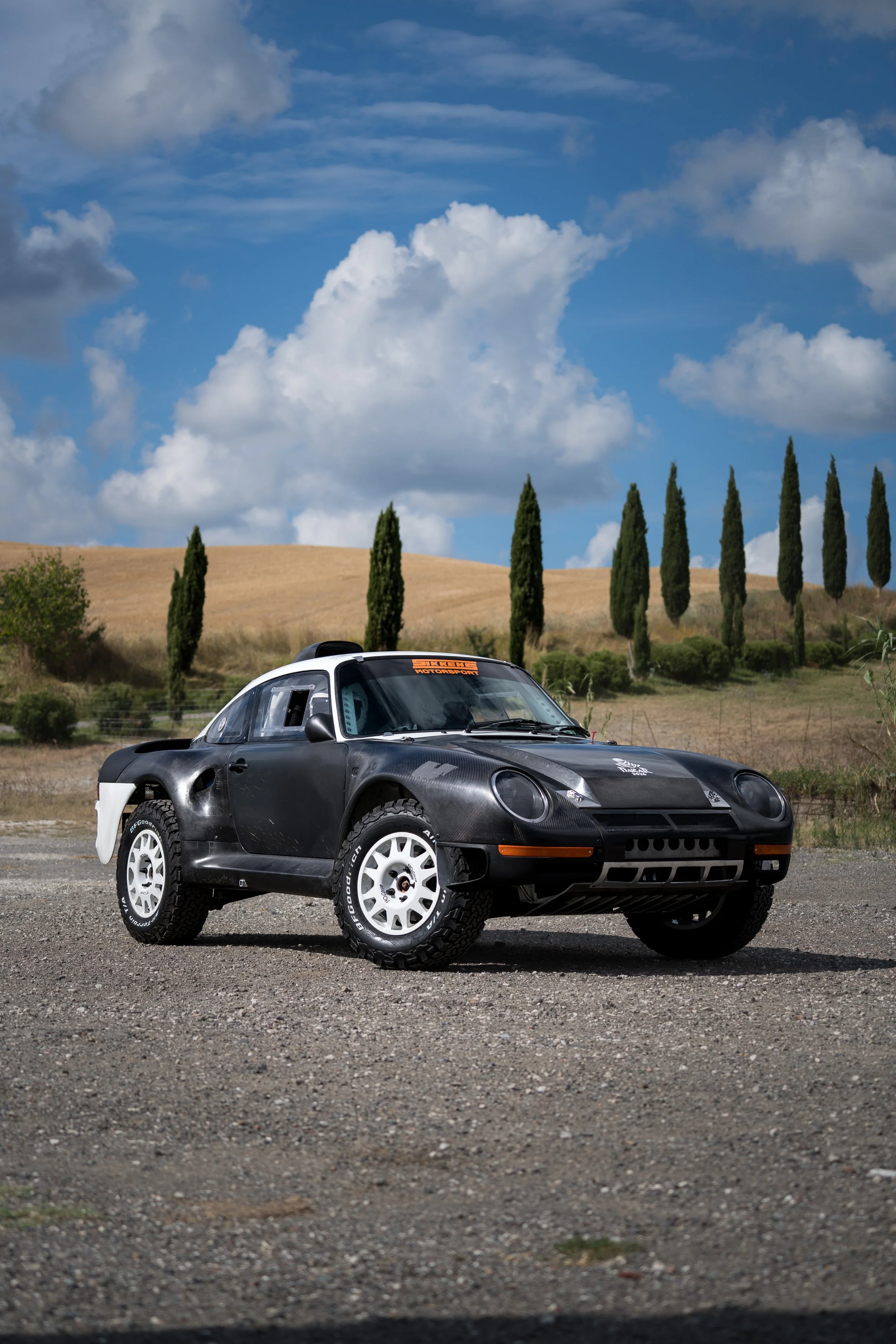 A black rally car with white wheels parked on a gravel surface with a scenic background of rolling hills, green trees, and a partly cloudy sky.