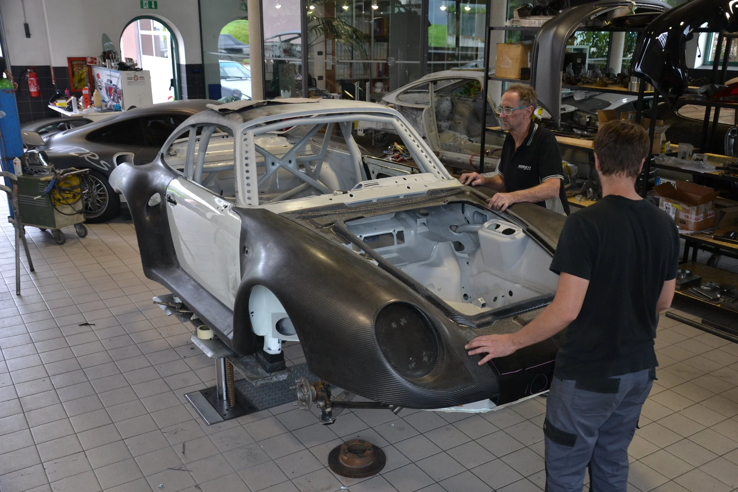 Two men work on a partially assembled race car in a workshop. The car has a carbon fiber body and a roll cage, with the interior and engine bay exposed.