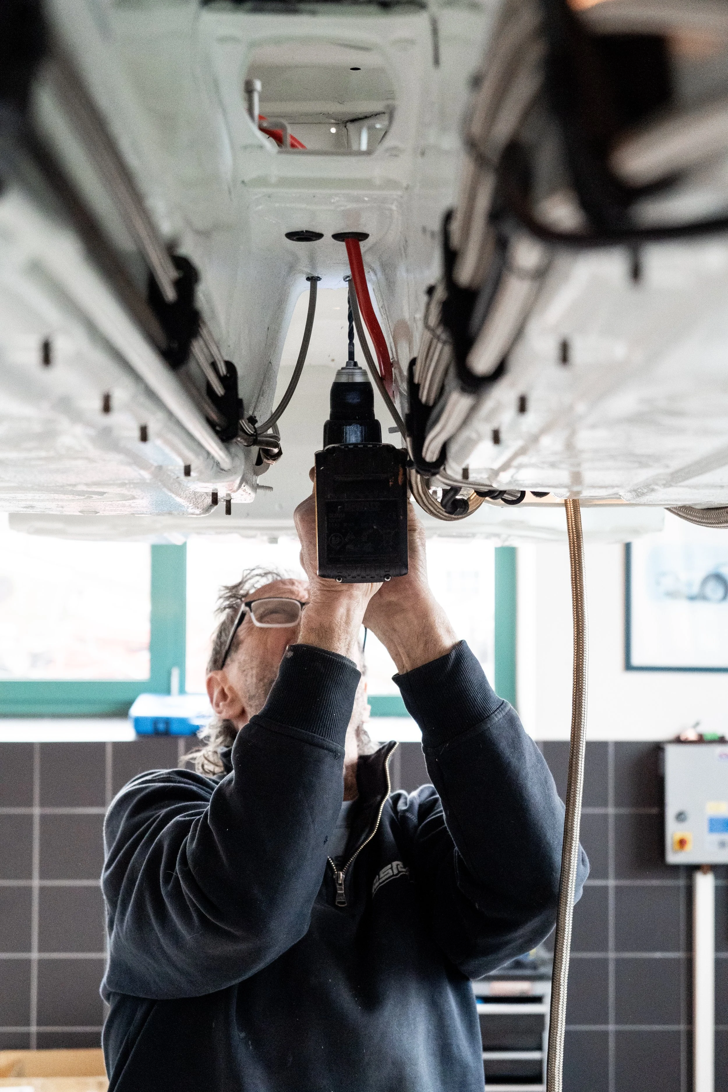 A man working underneath a vehicle, using a power tool, in a garage or workshop.