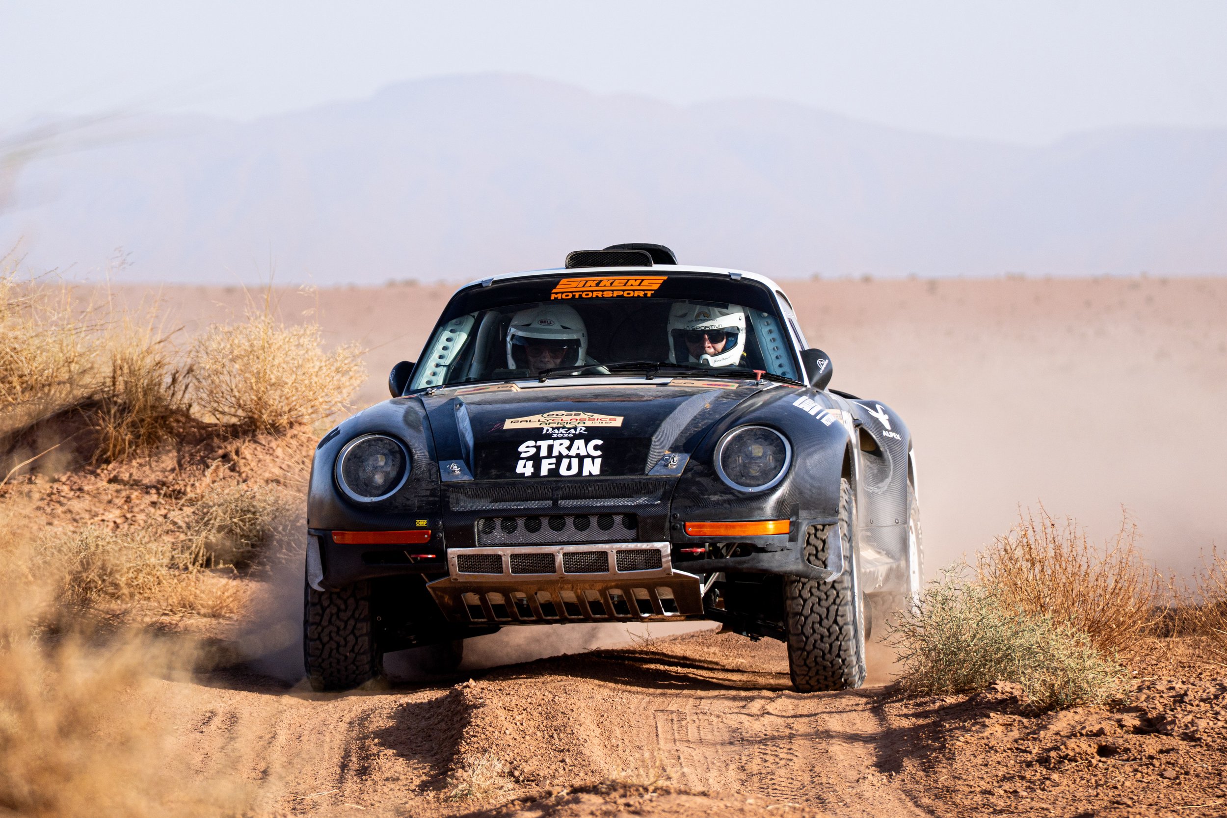 A black rally car driving on a dirt desert trail with dust kicking up behind it, two drivers wearing helmets visible through the windshield, and desert shrubs on either side of the trail.