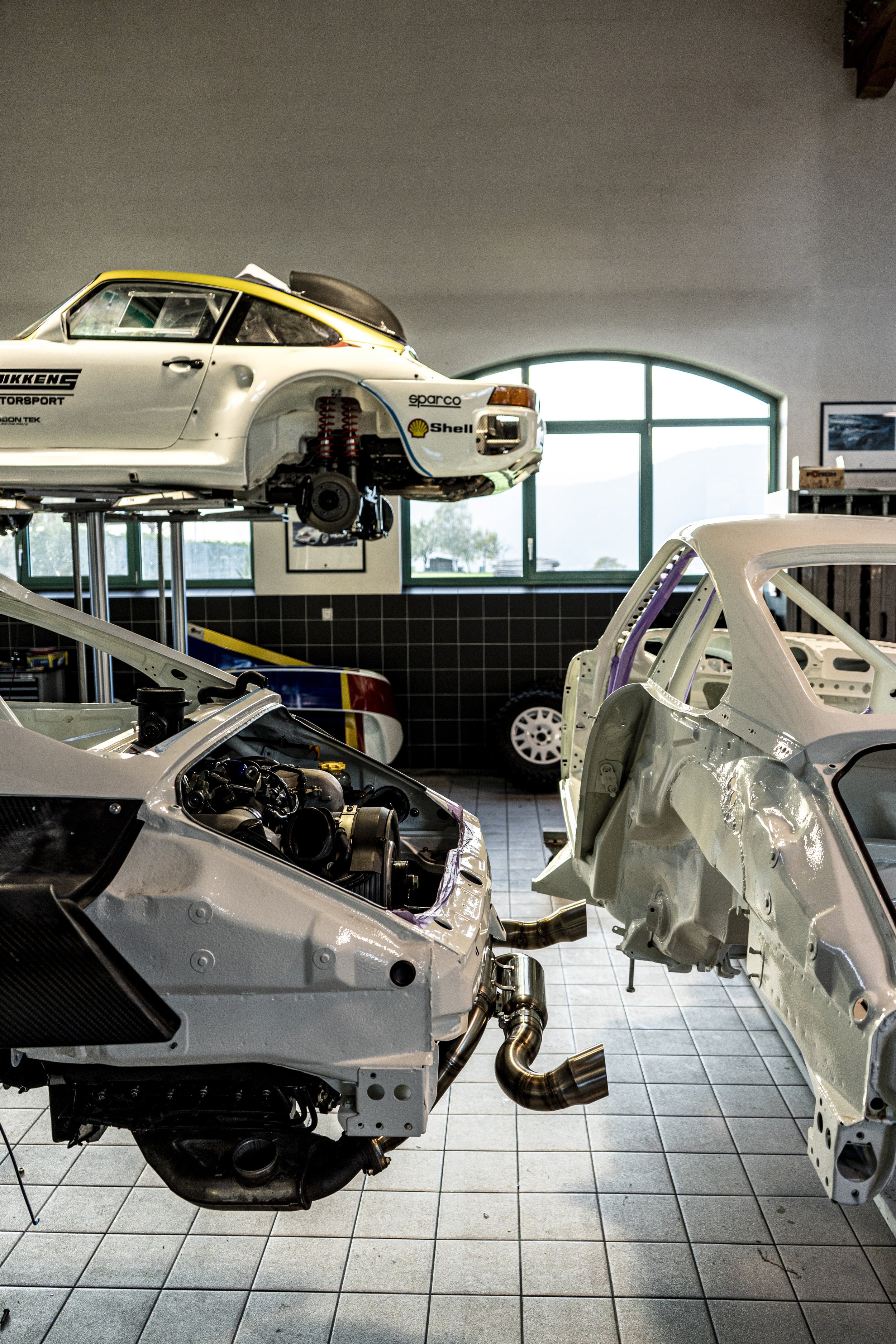 A garage with two partially assembled racing cars and a spare car wheel on the back wall.