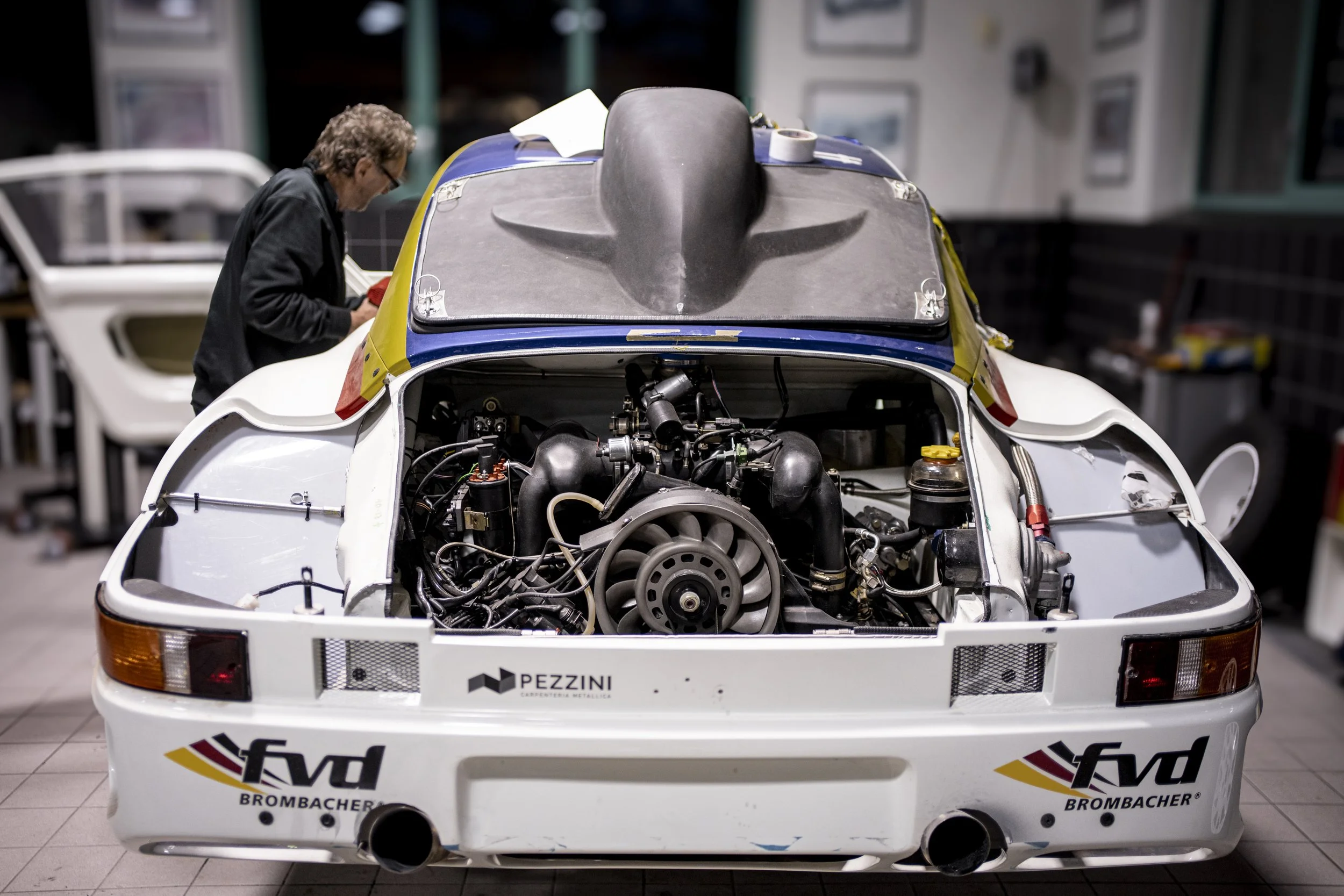 A racing car with its back cover removed, exposing the engine. A person is working on or examining something on the side of the car.