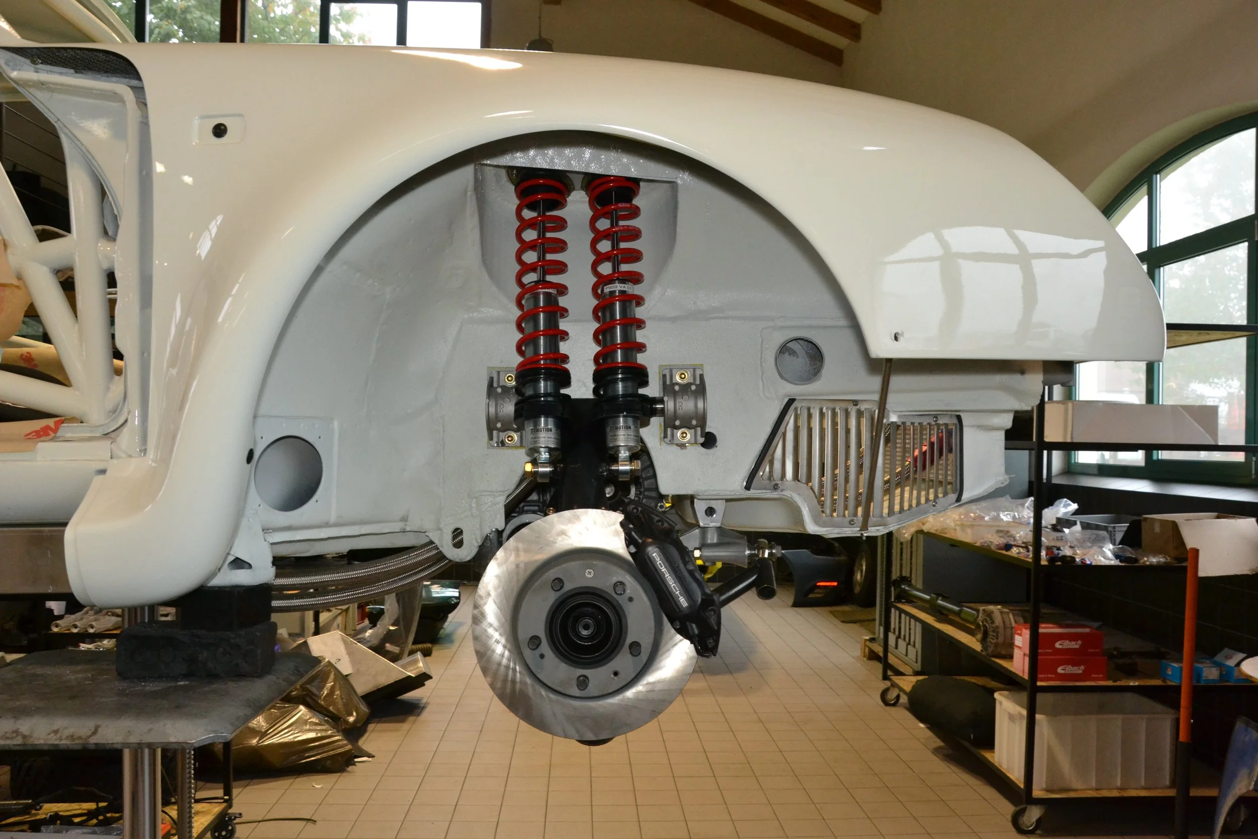 The image shows the front suspension and brake assembly of a race car, with red coil springs, a brake disc, and caliper, mounted in a partially assembled white chassis inside a workshop.