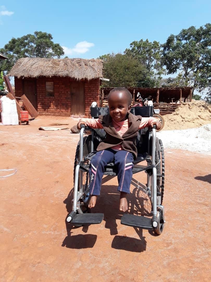 A smiling young boy sitting in a wheelchair outdoors on a dirt ground with a thatched-roof brick building and trees in the background.