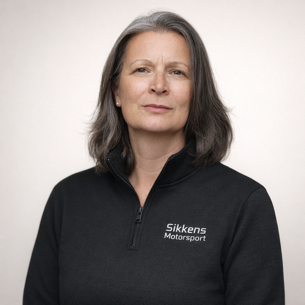 A middle-aged woman with shoulder-length gray hair wearing a black zip-up jacket with "Sikkens Motorsport" written on it, standing against a plain white background.
