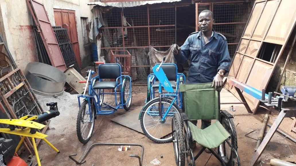 A man in a dark blue shirt with gloves stands in an outdoor workshop area with various wheelchairs and equipment around him.