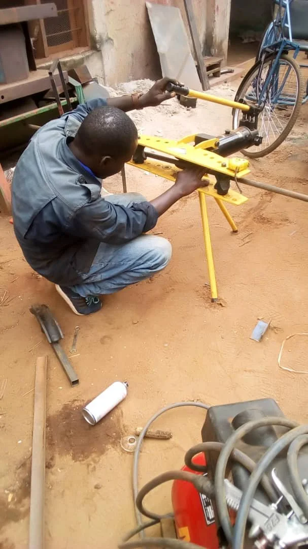 A man is crouched down working on a yellow metal device outdoors, with a bicycle in the background and tools like a welding machine and metal pieces on the ground.