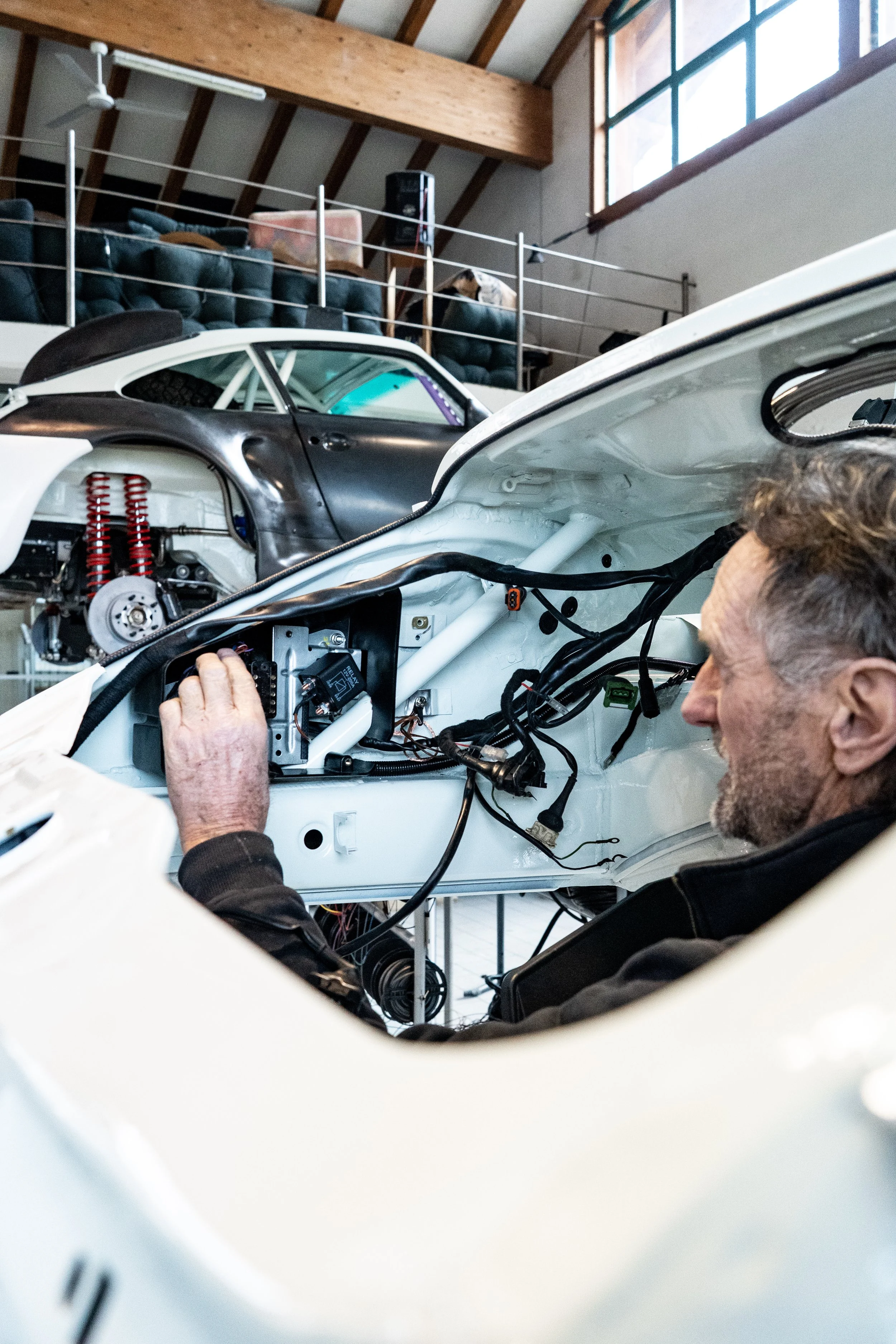 A man working inside a race car, with the car's interior exposed, in a garage or workshop with a high ceiling and large windows