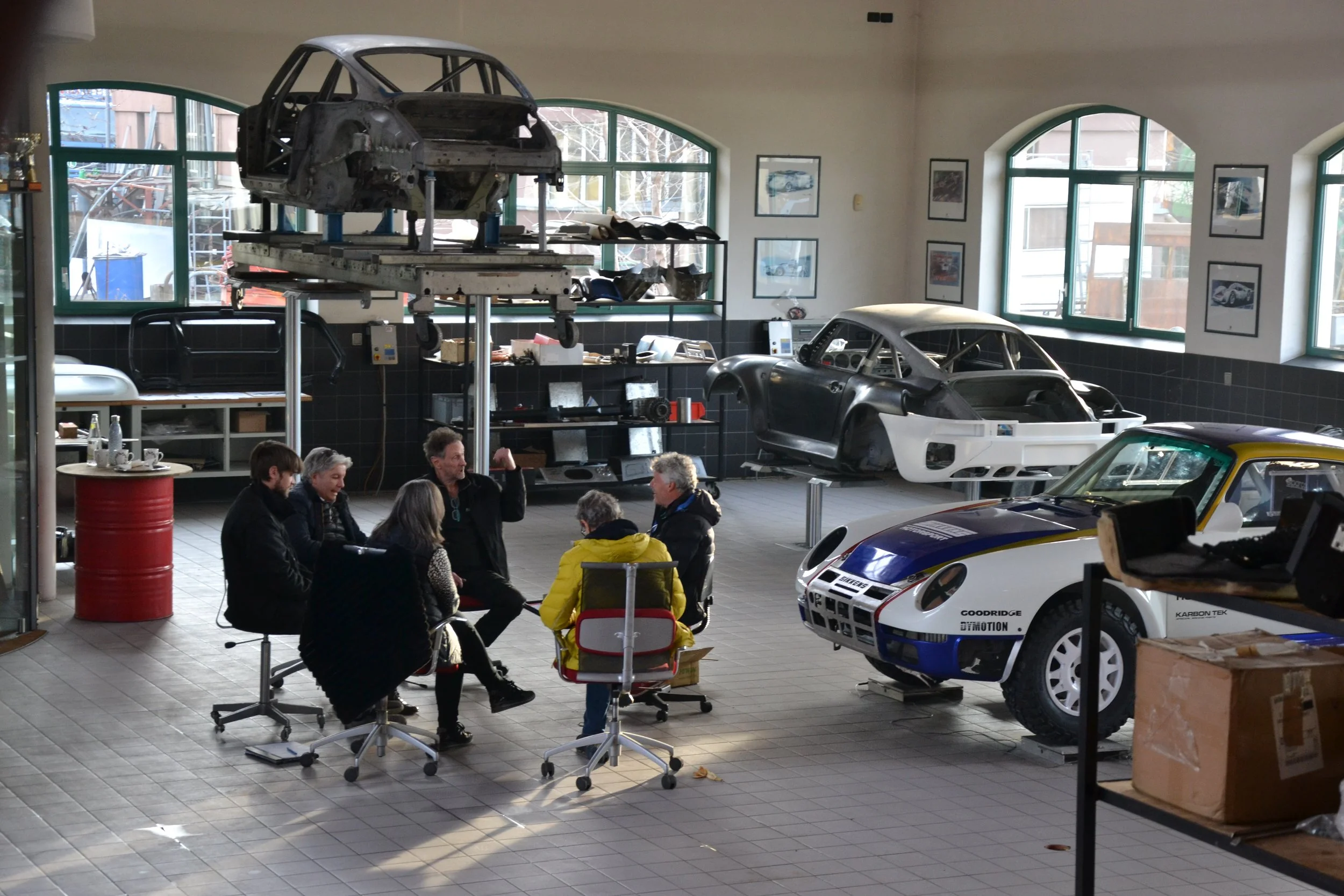 Group of people sitting and talking inside a workshop with race car bodies and chassis in the background.