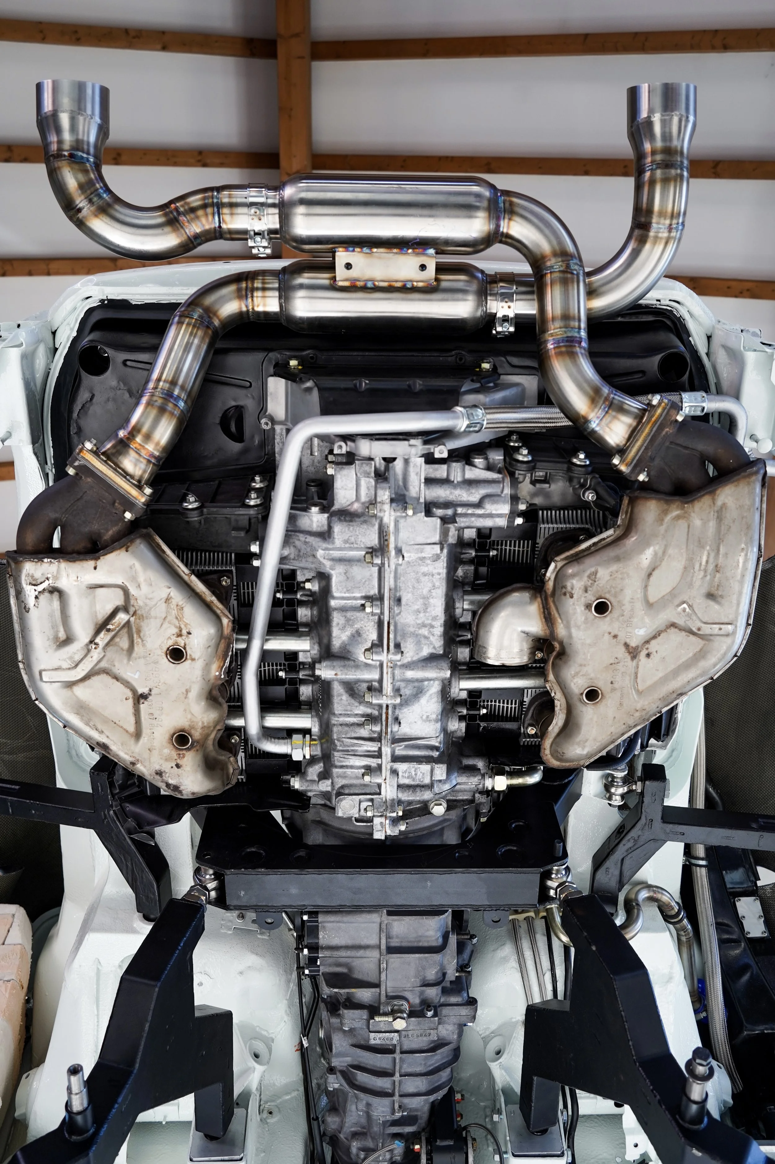 Close-up of a car engine with custom exhaust headers installed, showing metallic pipes and engine components inside a garage.