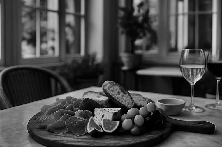 A cheese and charcuterie board with assorted bread, lemon wedges, grapes, and sliced meats on a round wooden platter in a cozy room with large windows, wine glasses, and a potted plant in the background.