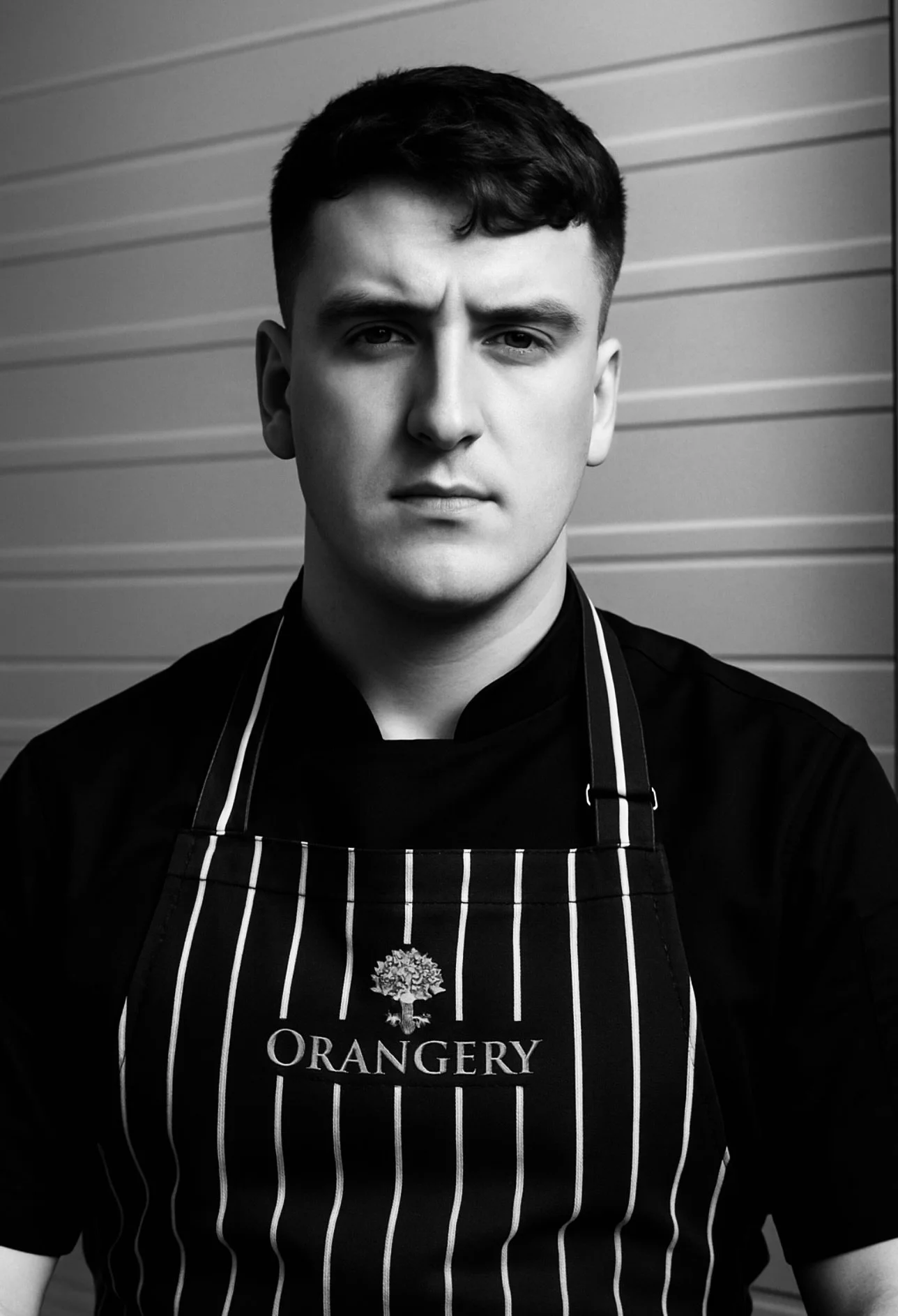 Black and white photo of a young man wearing an apron with the word 'O R A N G E R Y' and a tree logo on it, standing in front of a horizontal siding wall.