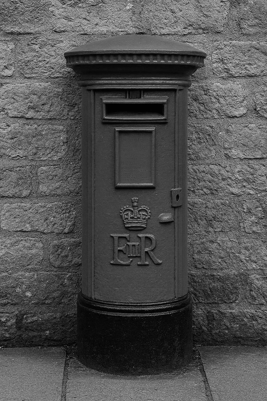 A vintage black metal mailbox with the royal crown and initials 'E R' on its front, mounted on a sidewalk against a stone wall.