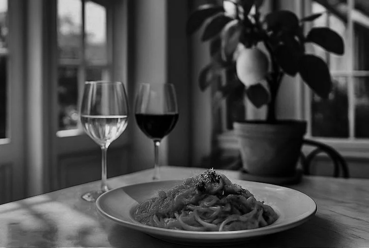 A plate of pasta with sauce, two glasses of wine (one white, one red), and a potted plant on a wooden table near a window with sunlight.