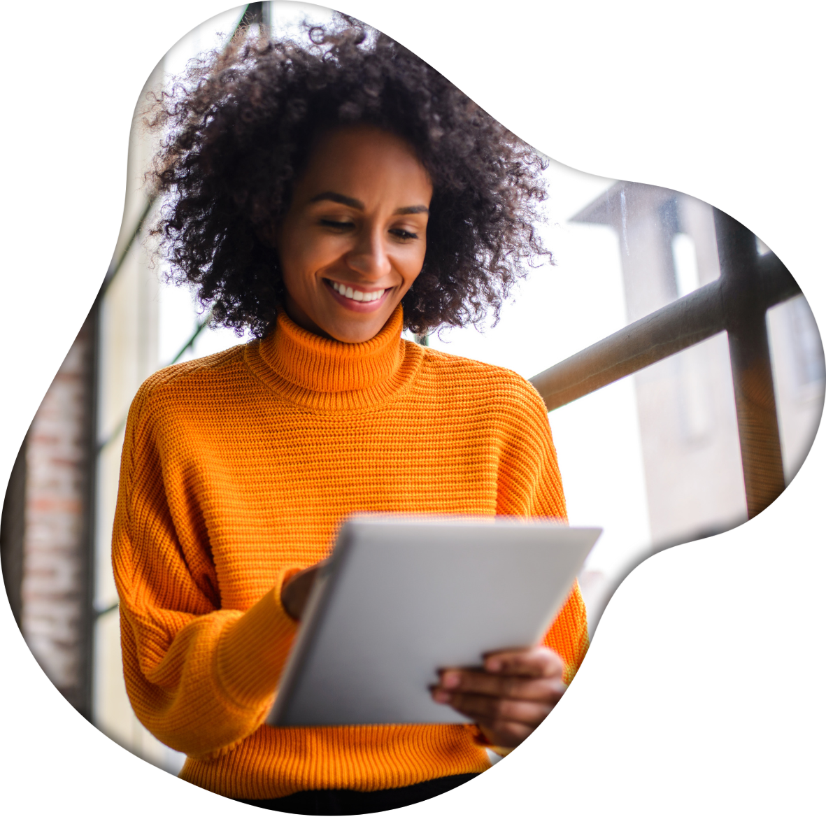 A woman with curly hair wearing an orange sweater, smiling while looking at a tablet, indoors near a window.
