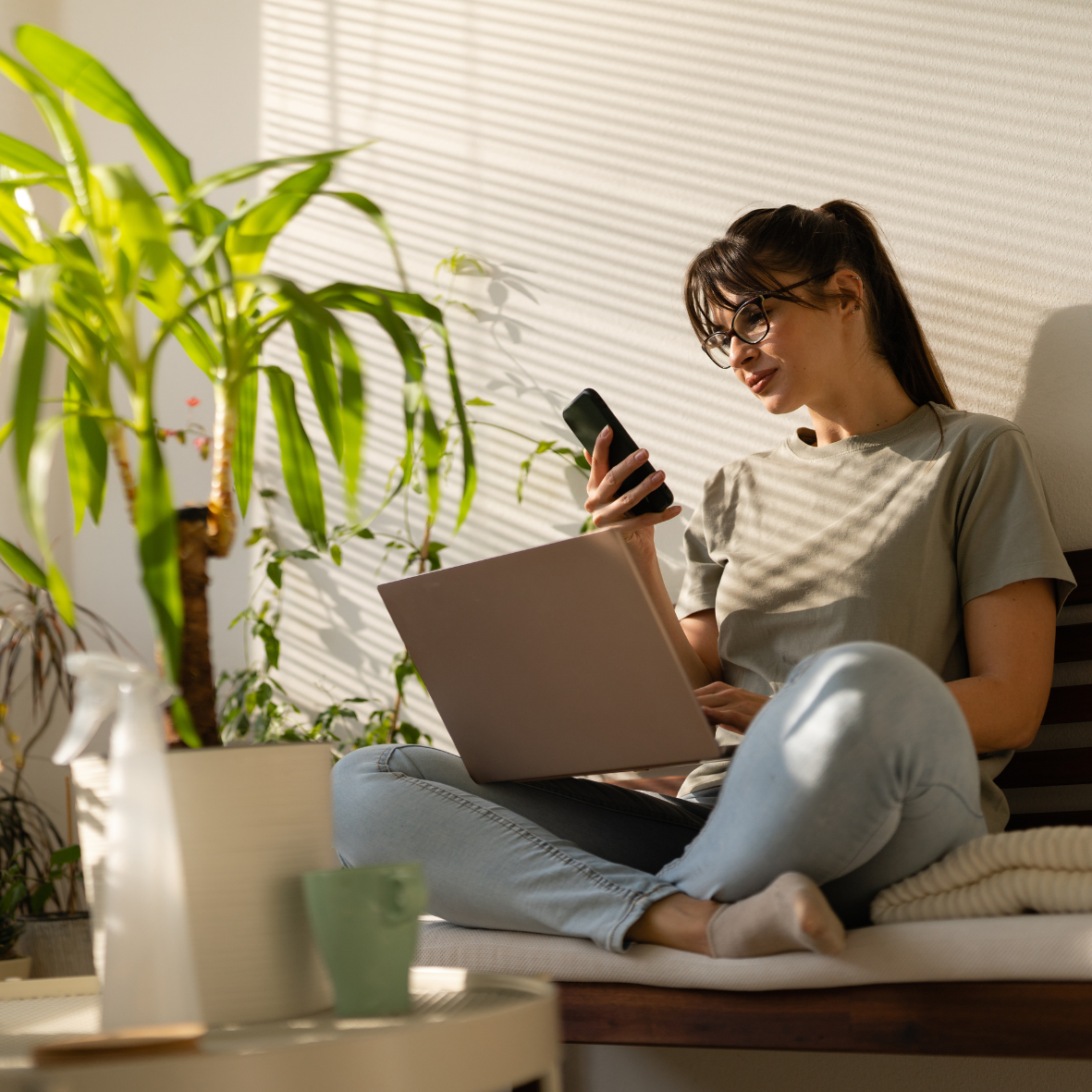 Young woman sitting on a couch with a laptop on her lap, using a smartphone, surrounded by houseplants with sunlight and shadows creating patterns on the wall.
