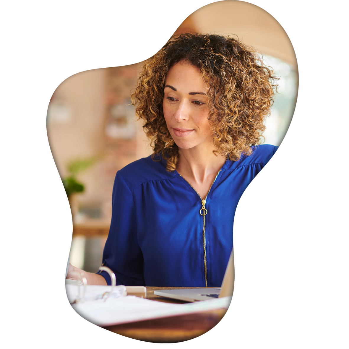 A woman with curly hair wearing a blue top, looking down at a desk with papers and a laptop, in a room with a brick wall and blurred background.