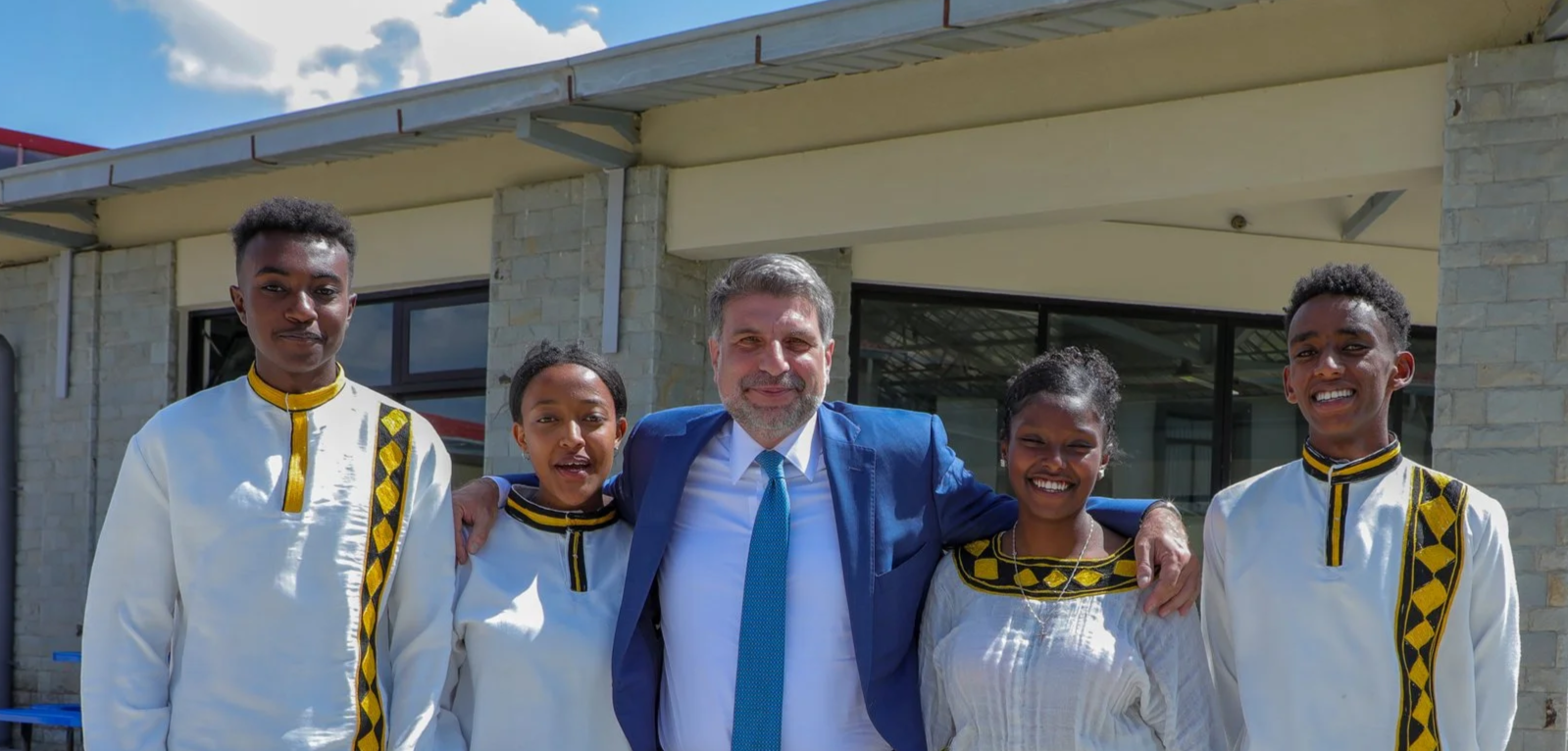 Group of five people standing outside, smiling, with a building in the background. Four are wearing traditional clothing with yellow and black embroidery. A man in a blue suit stands in the center with his arms around the others.